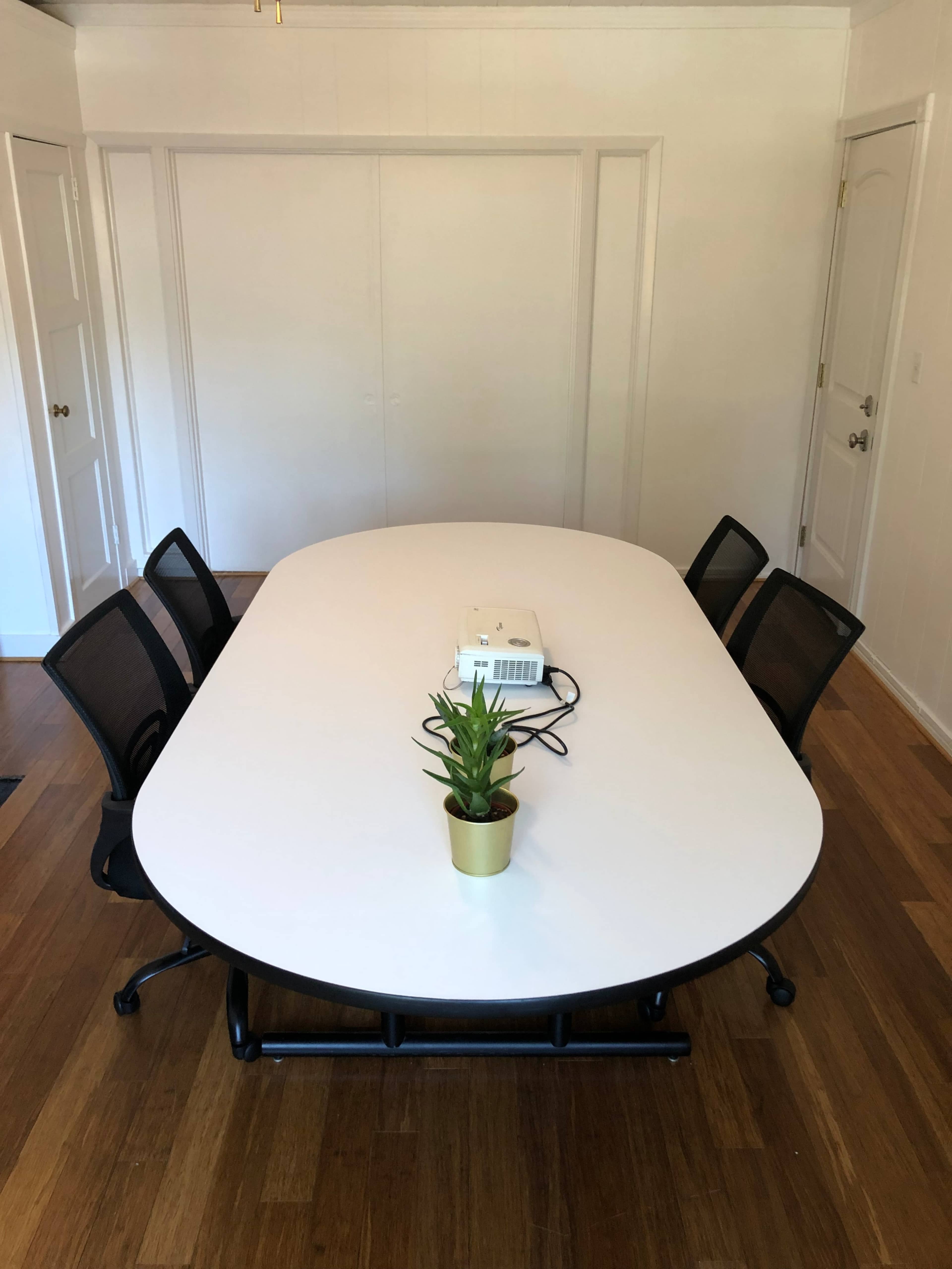 A large white conference table with a small plant centerpiece is surrounded by five black office chairs in a bright, minimalist room.