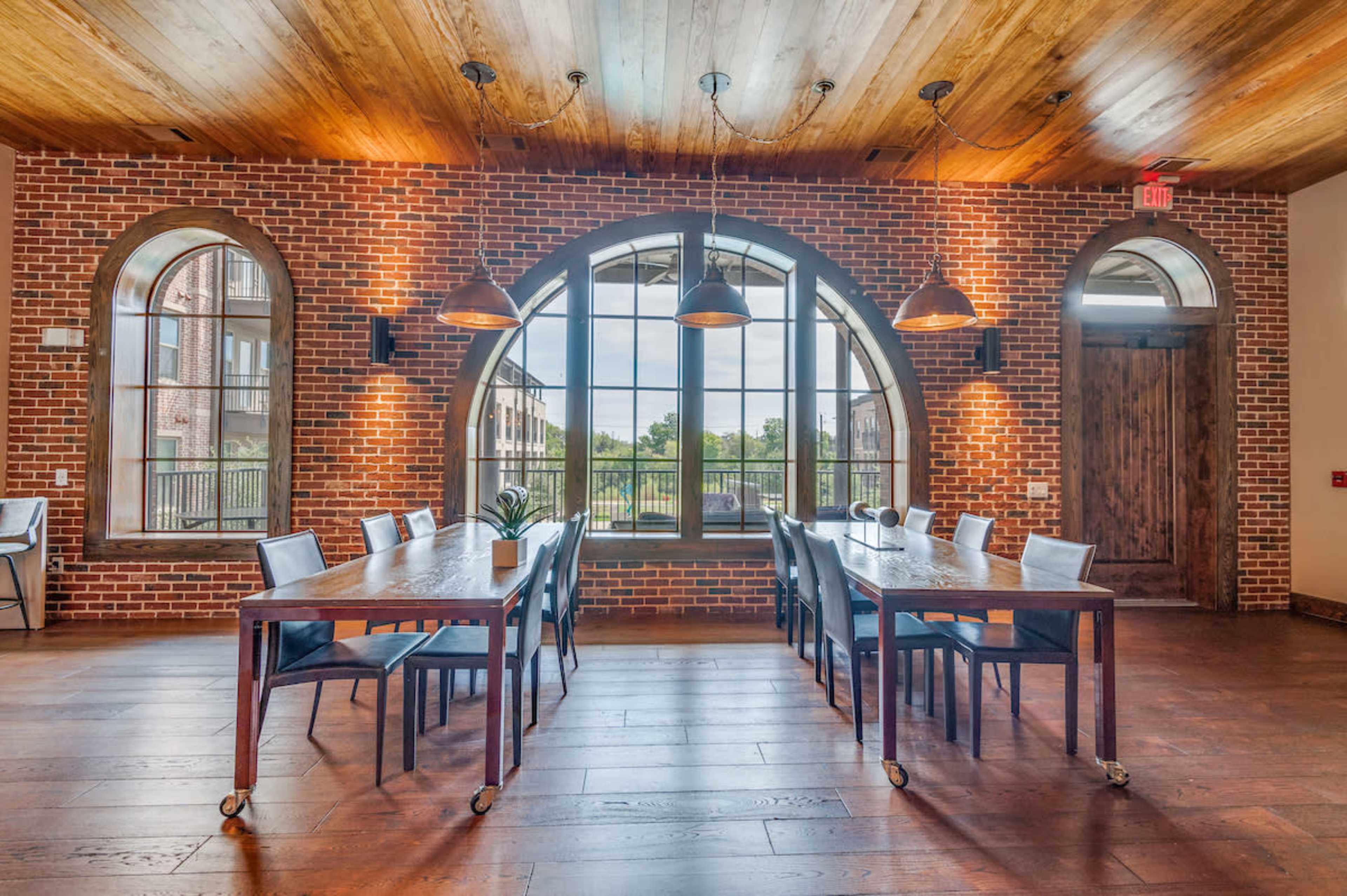 A spacious dining area features wooden tables and chairs under pendant lights, with large arched windows and a brick wall backdrop.
