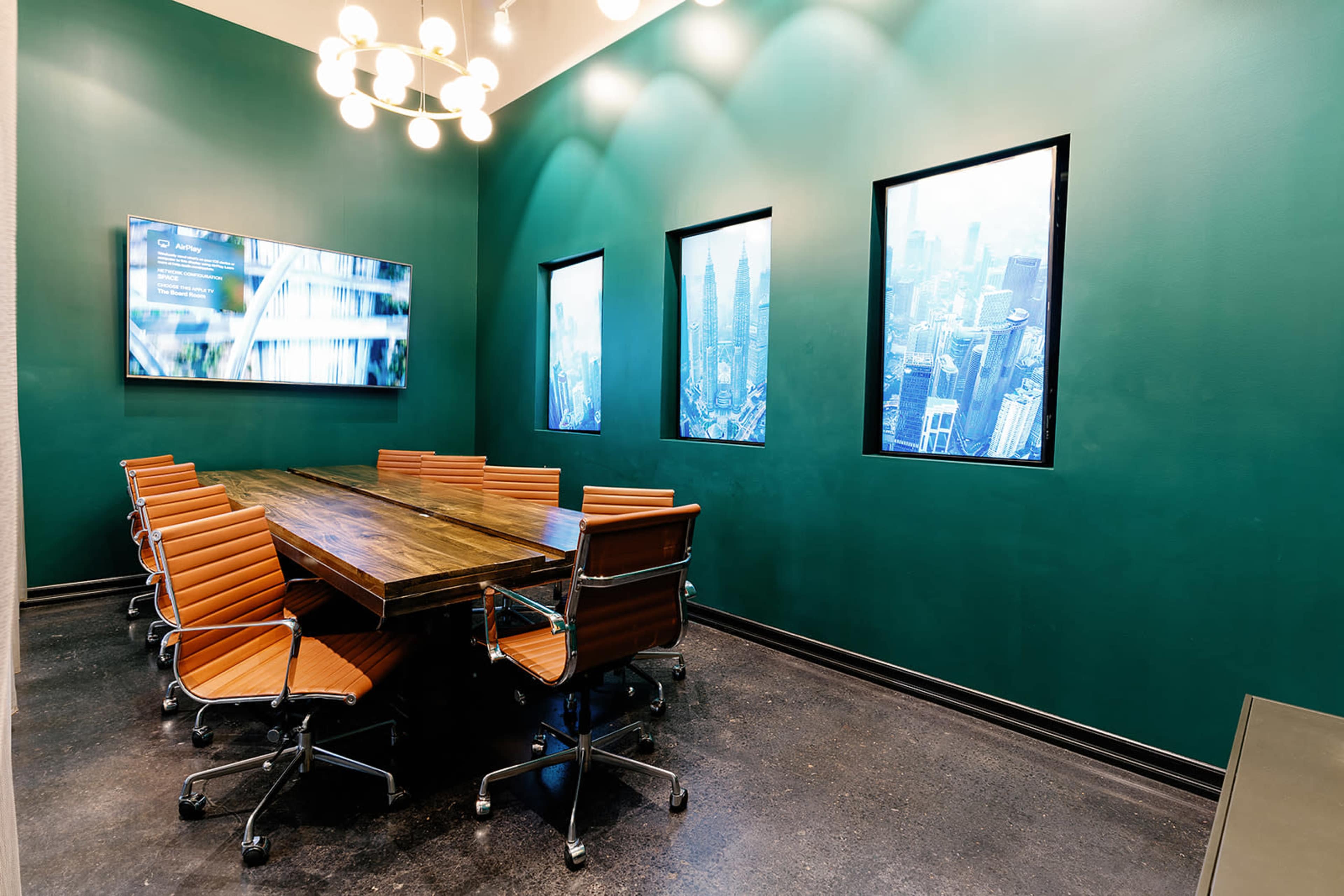 A modern conference room features a long wooden table surrounded by orange office chairs, with green walls and large windows displaying cityscape images.