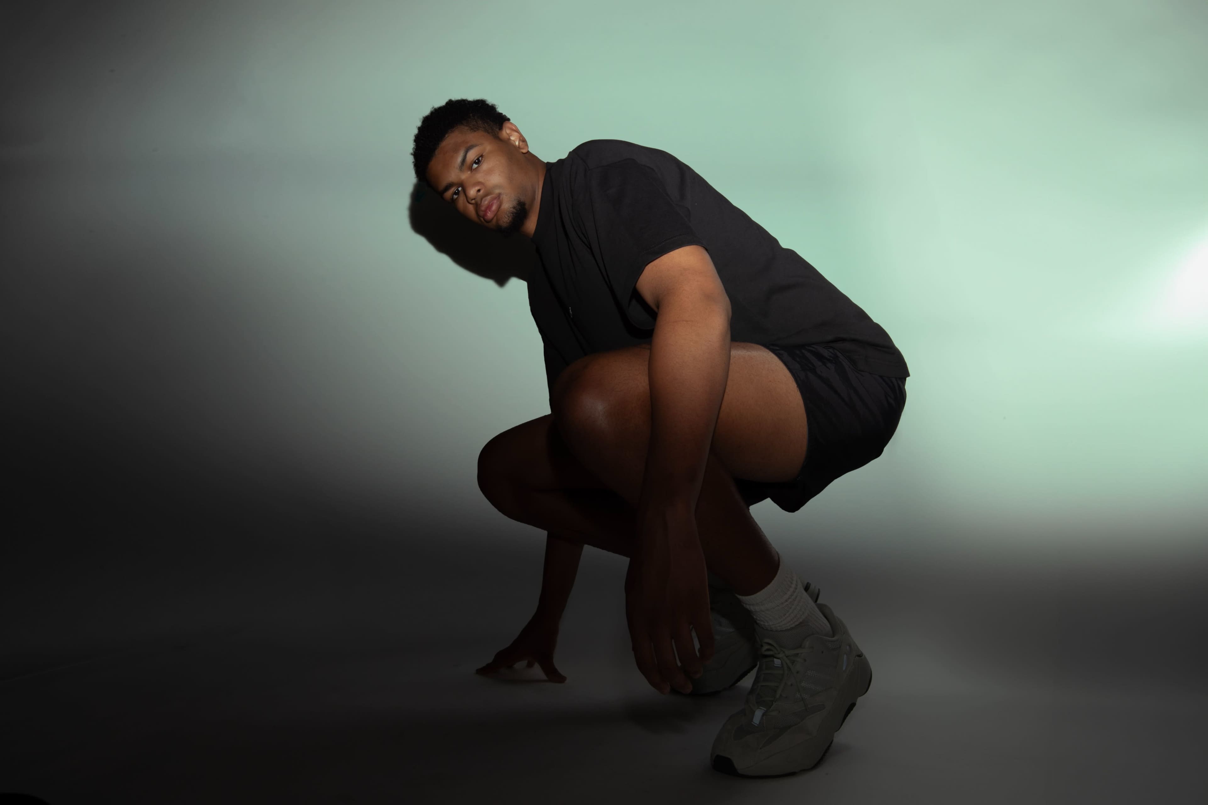 A man in athletic wear is crouching on a gray backdrop with soft lighting.