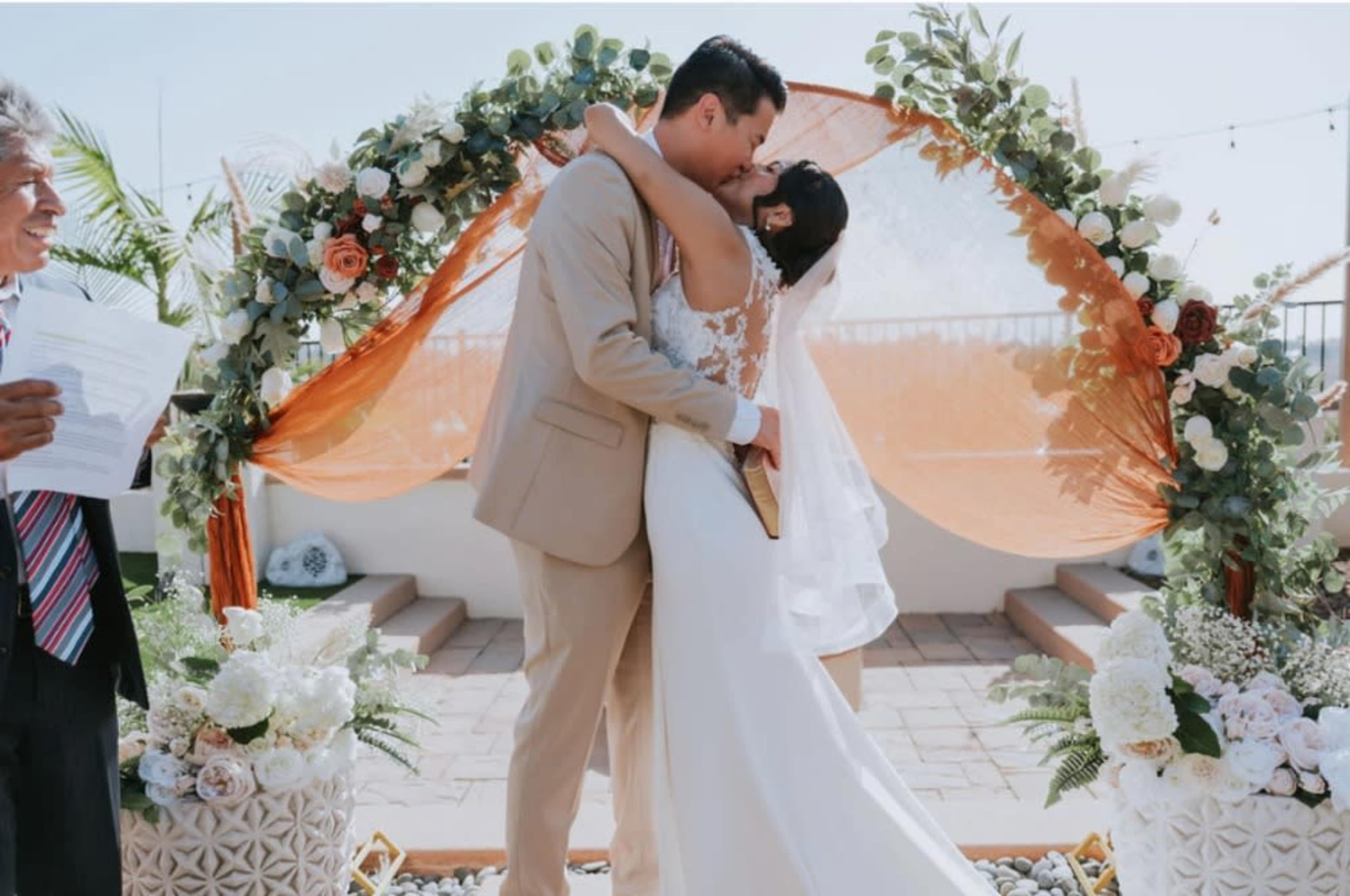 A couple embraces in a kiss under a floral arch during their wedding ceremony.
