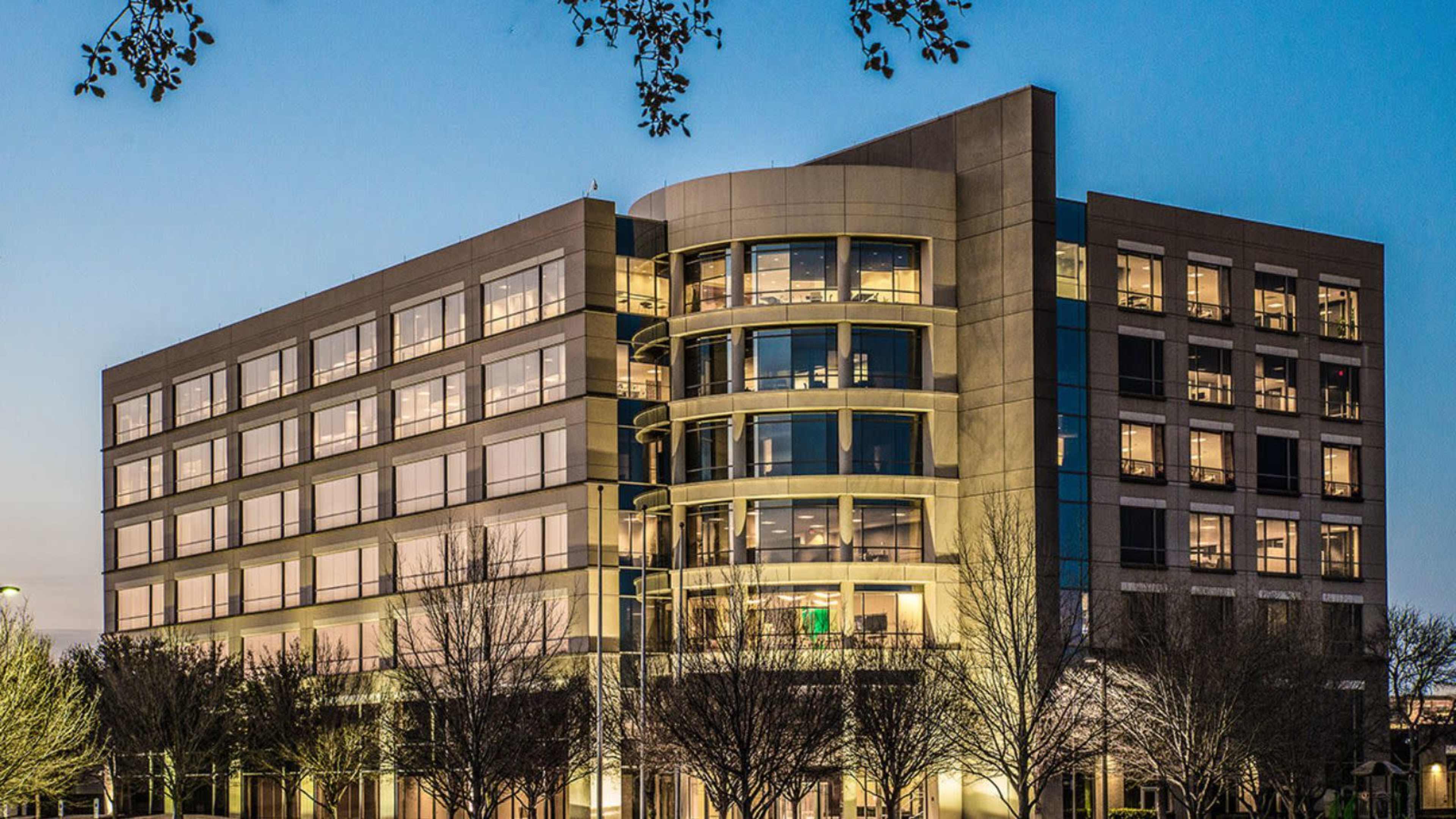 The image shows a modern, multi-story office building illuminated at dusk, with large windows and a curved facade.