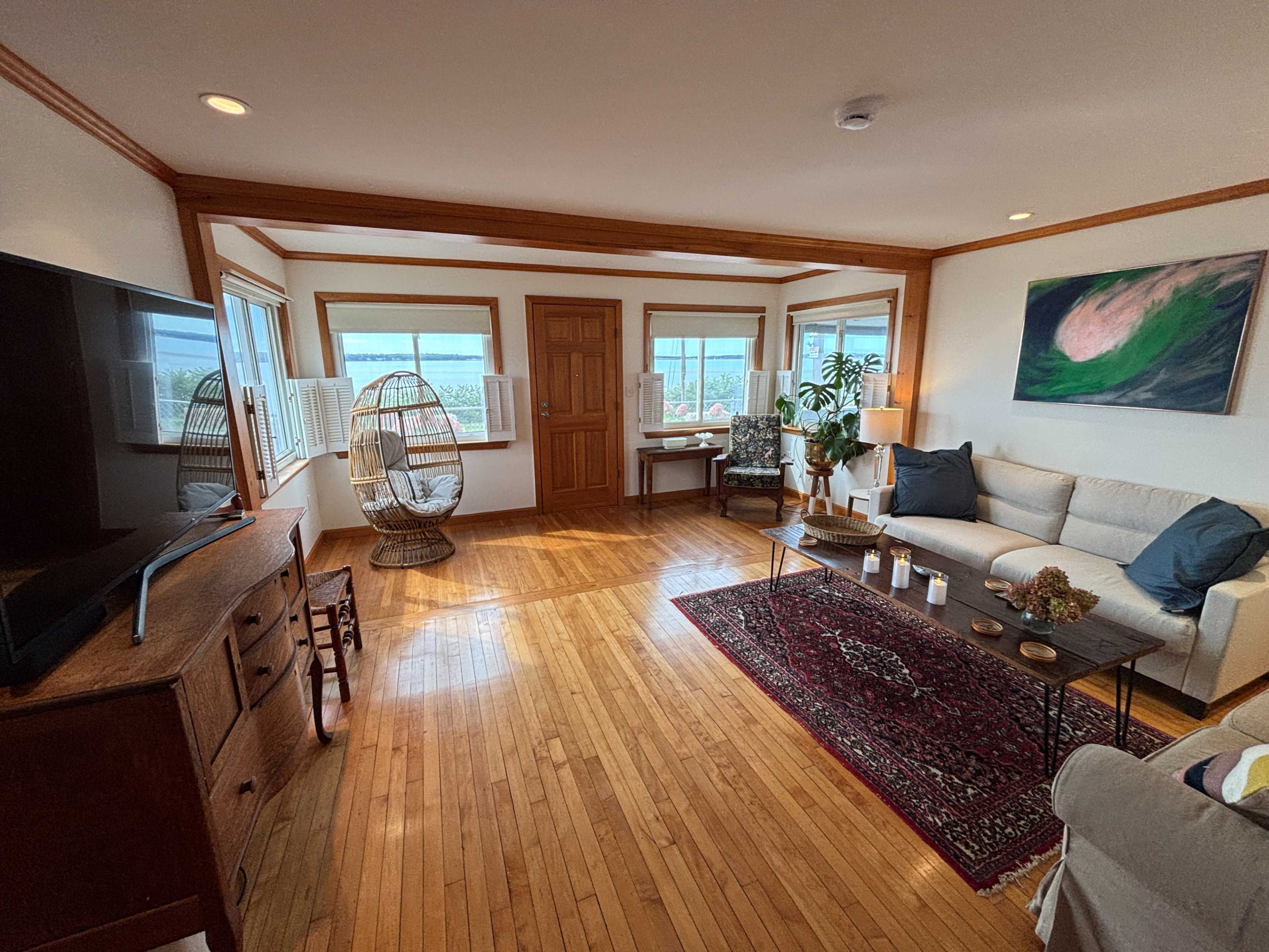 The image shows a cozy living room with hardwood floors, a sofa, a coffee table, a chair with a floor lamp, and large windows providing a view of the outside.