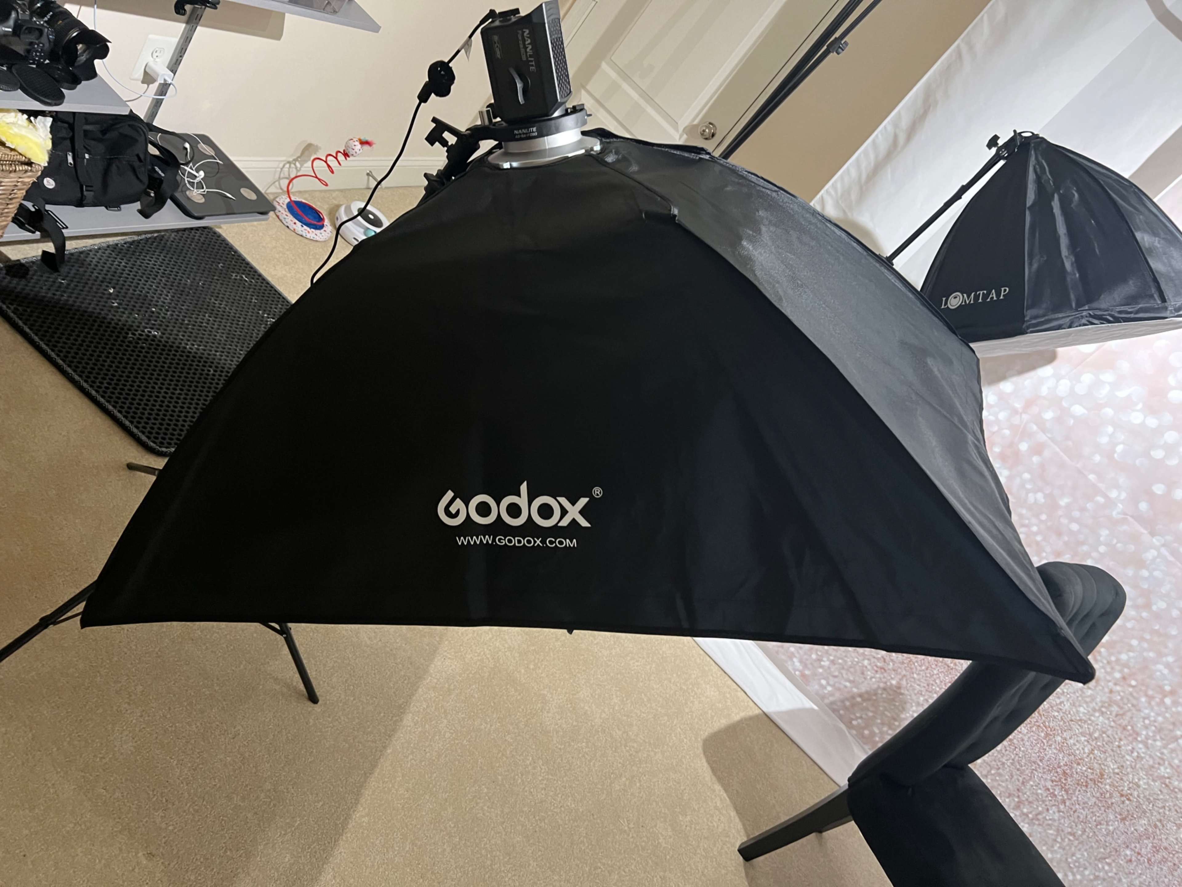 A black Godox softbox positioned in a photography studio setting with a tripod and other equipment visible in the background.