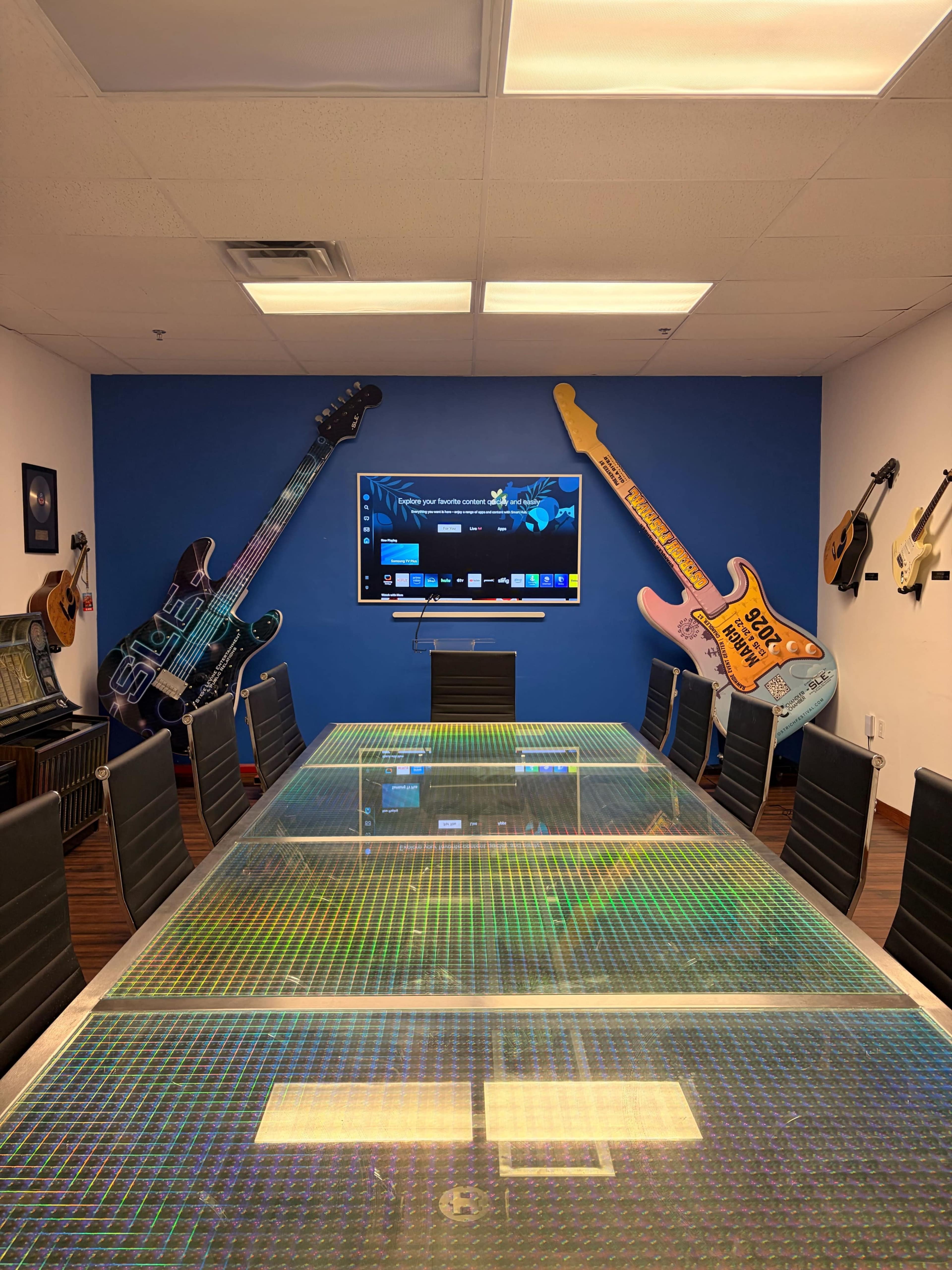 A conference room features a long glass table surrounded by black chairs, with large guitars mounted on the blue wall and a television screen at one end.