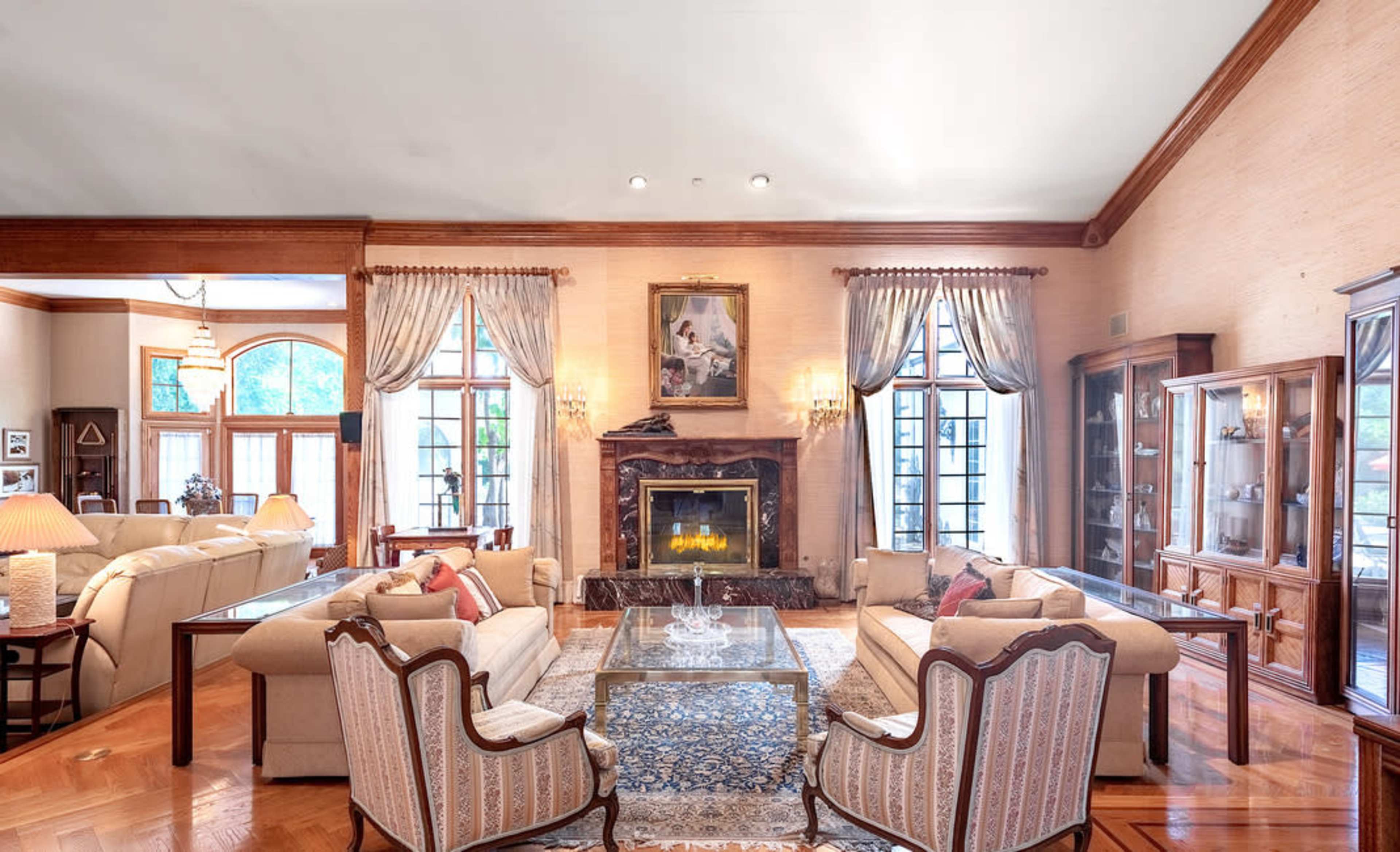 The image shows an elegant living room with large windows, a fireplace, and stylish furniture arranged around a glass coffee table.