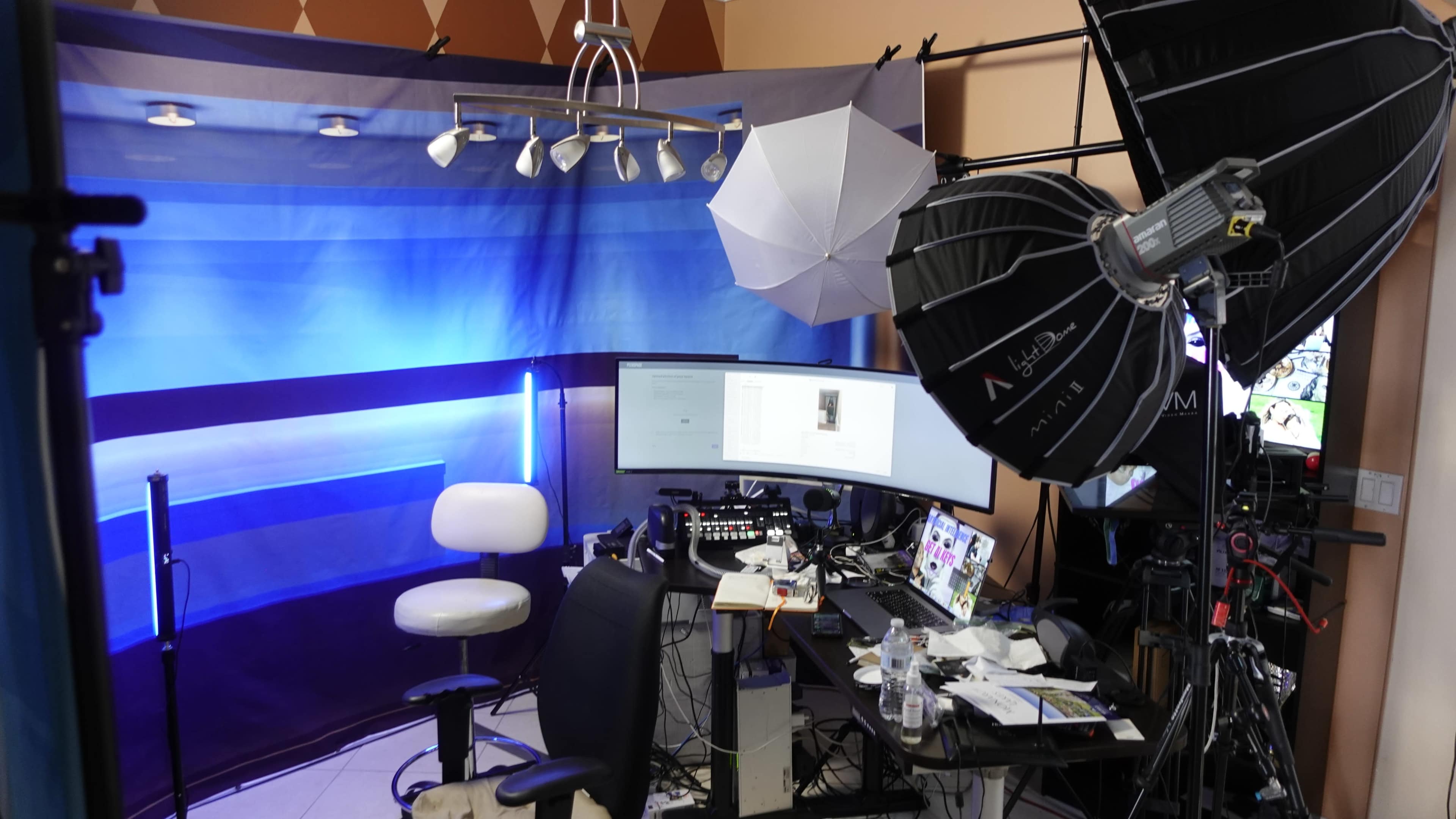 A video production setup features multiple lighting sources, a desk with dual monitors, and a blue backdrop.