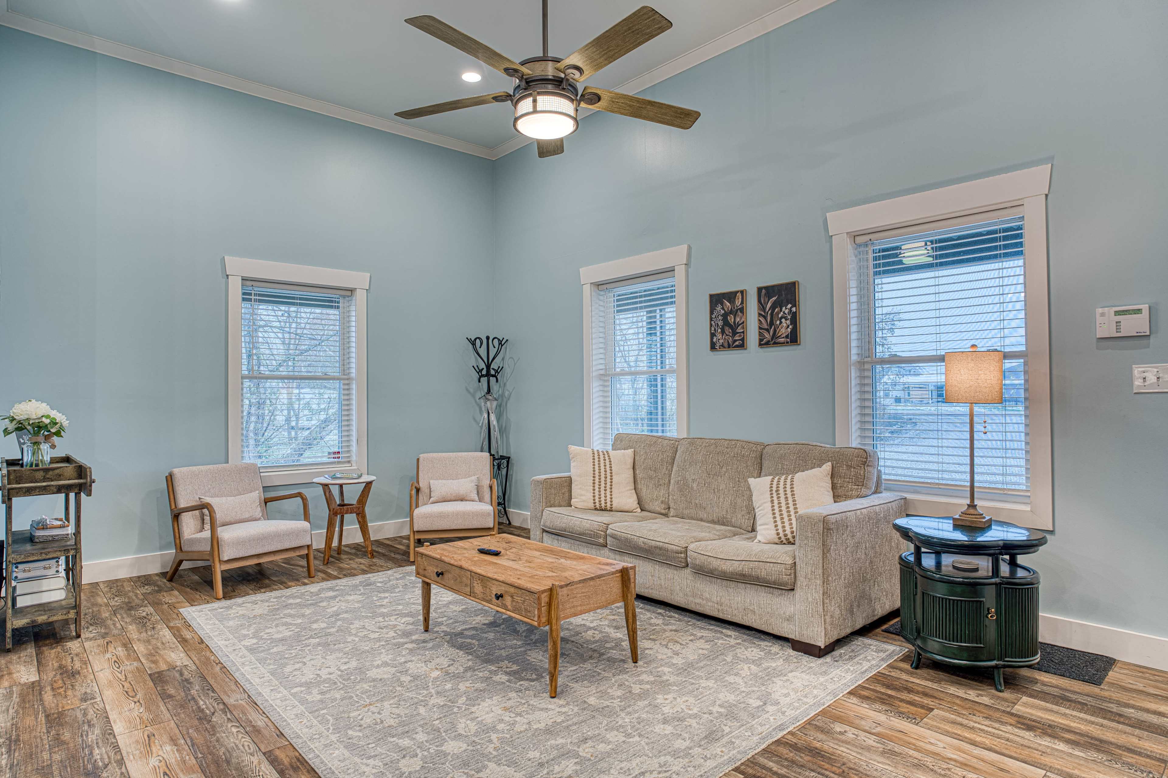The image shows a cozy living room with a light blue wall, a beige couch, two chairs, a wooden coffee table, and large windows.