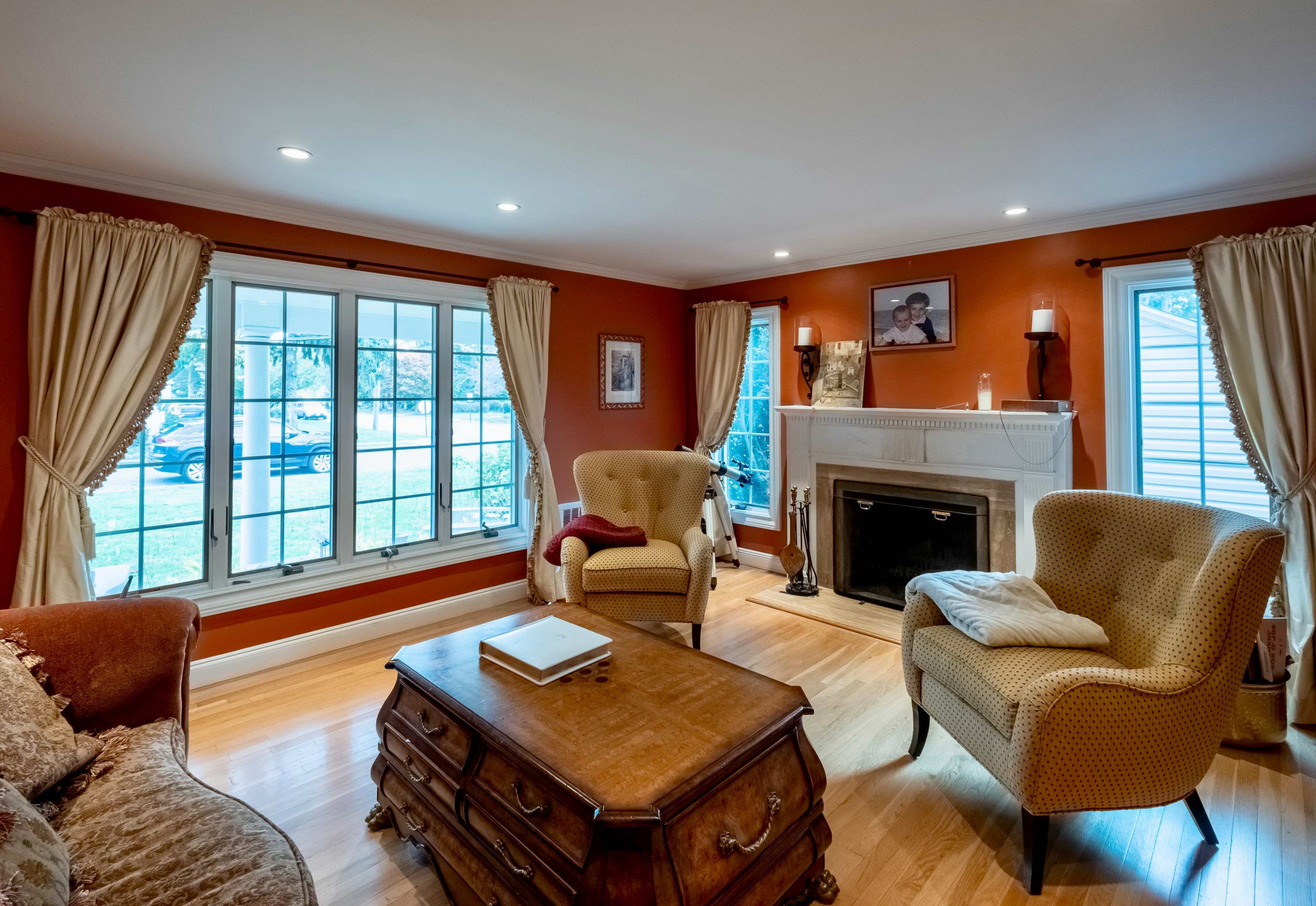 The room features a cozy seating area with two armchairs and a sofa, a wooden coffee table, and a fireplace against a wall adorned with windows and curtains.
