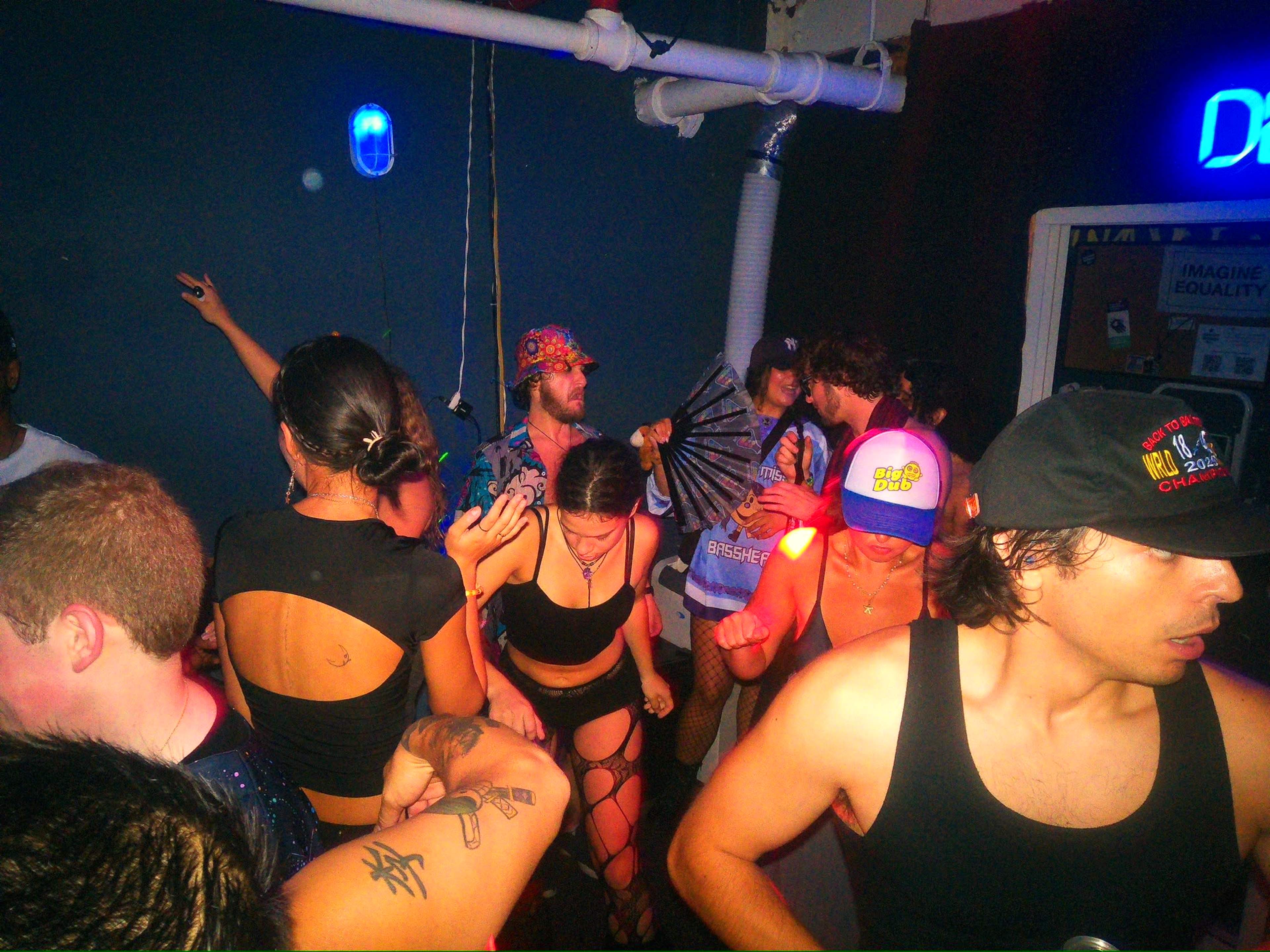 A group of people dance in a dimly lit room with blue lighting and various clothing styles.