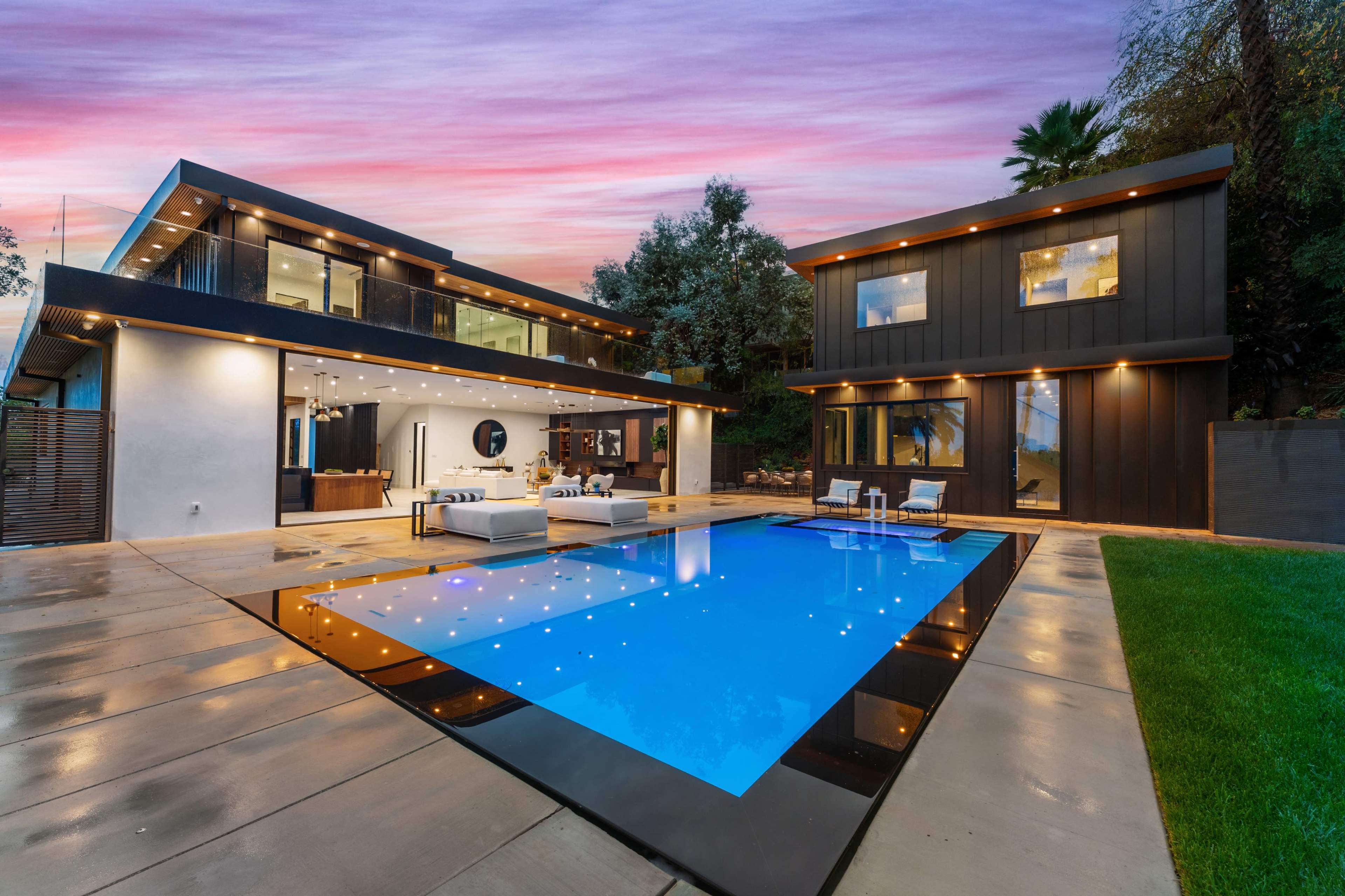 A modern home features two structures with large windows, a pool with a reflective surface, and outdoor seating areas set against a colorful sunset sky.
