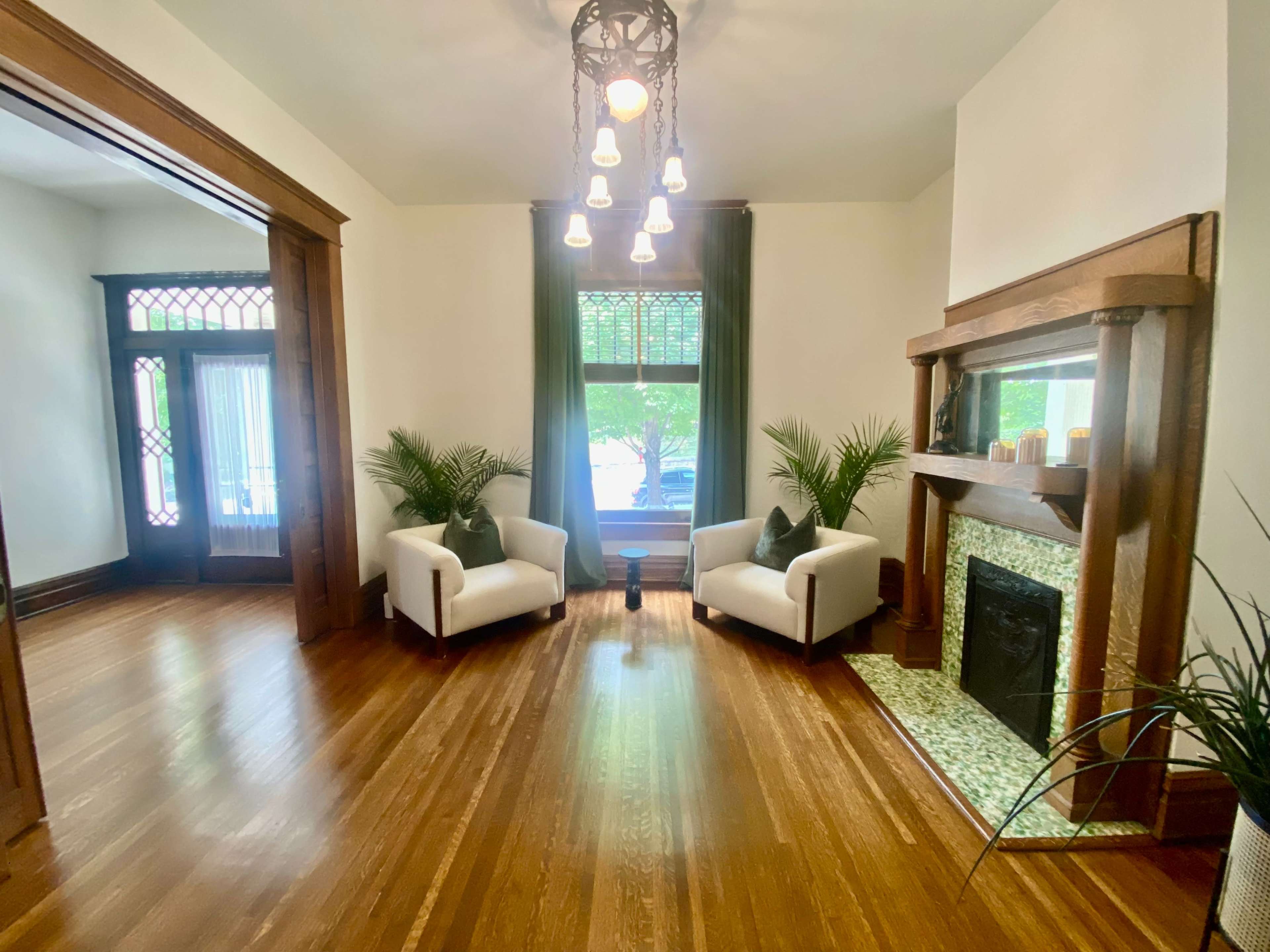 The image shows a cozy room with two white armchairs, a fireplace with a green tiled surround, and large windows framed by green curtains.