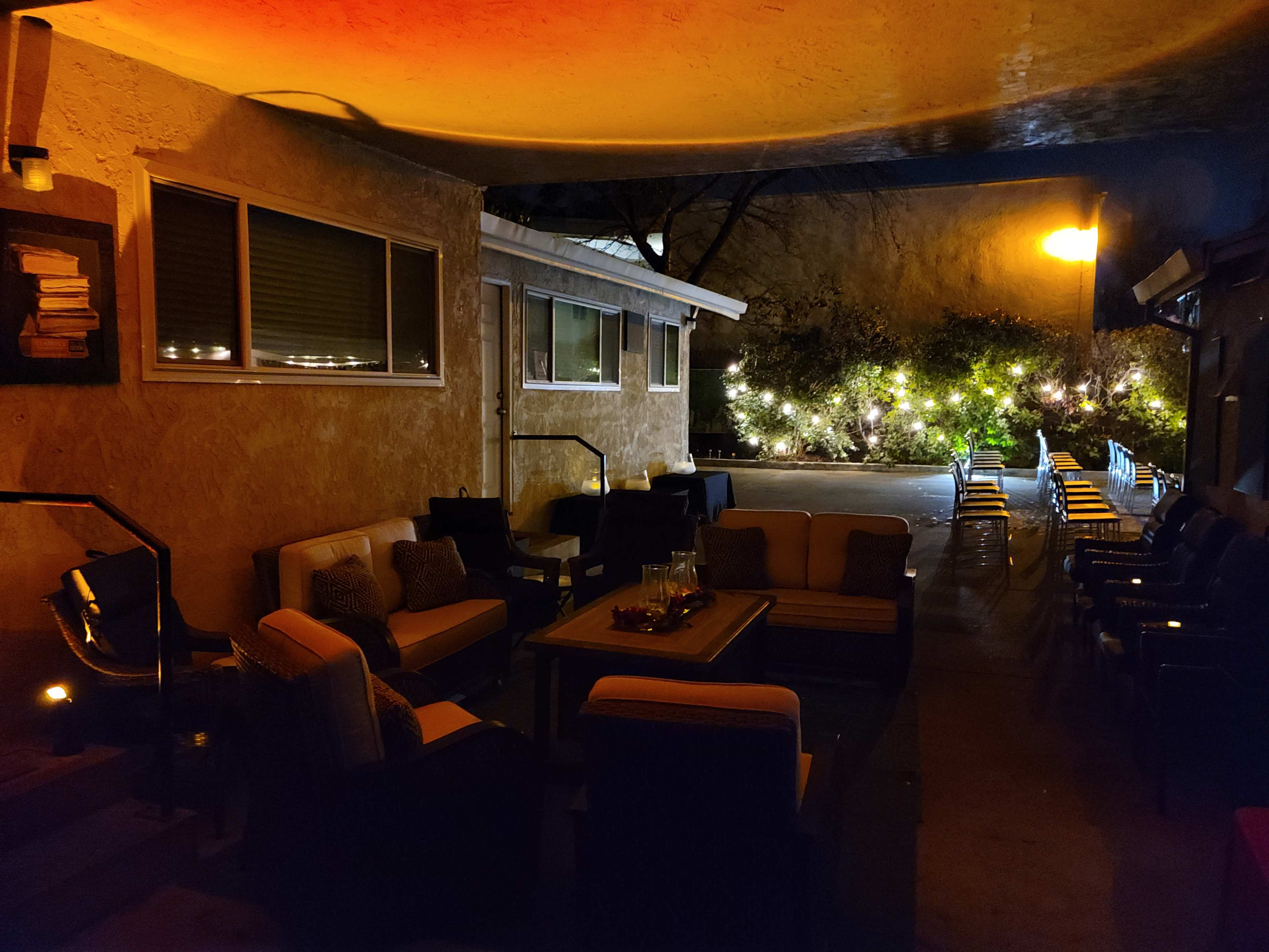 A dimly lit outdoor seating area with couches and chairs arranged around a central table, leading toward a pool area illuminated by lights.