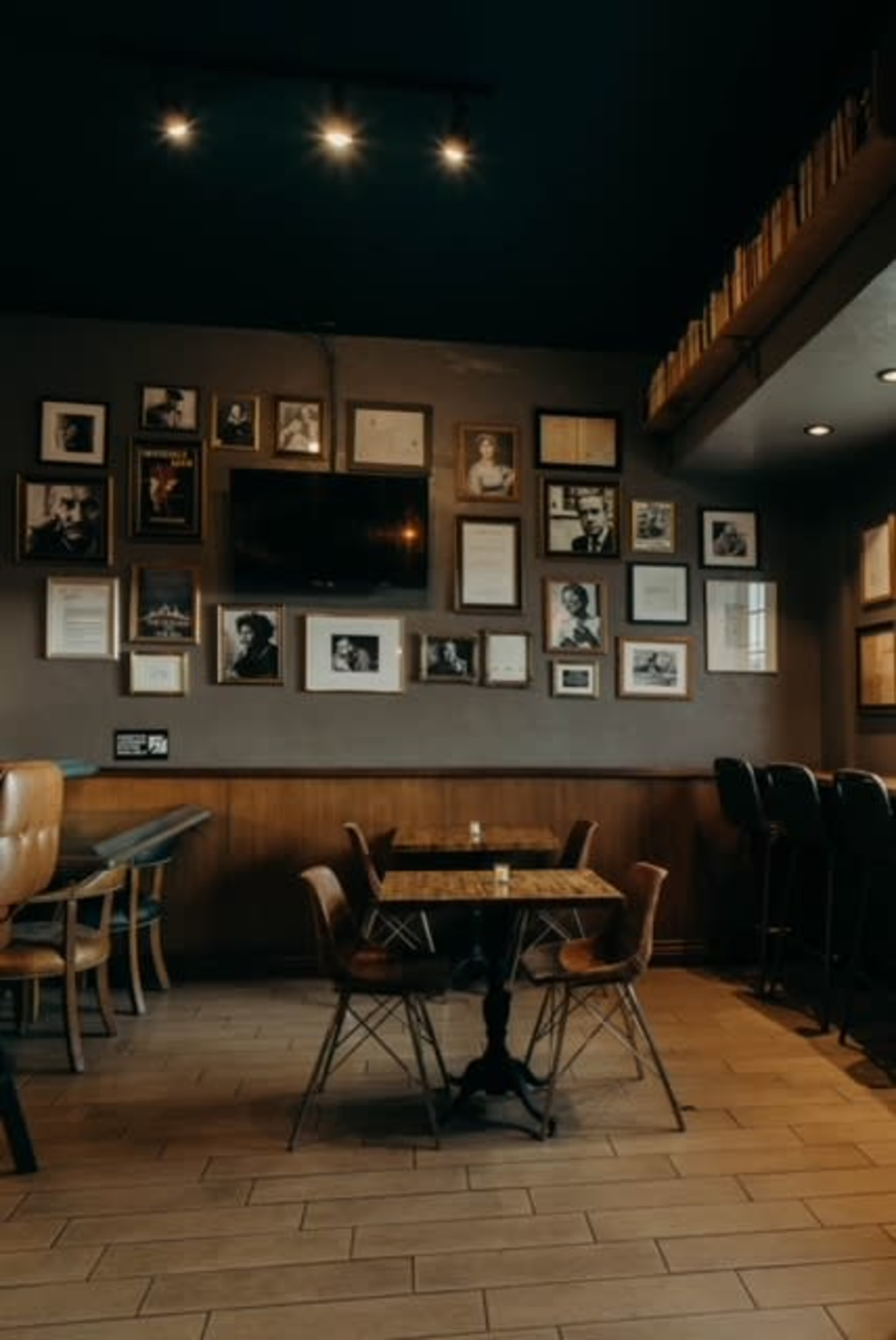 The image shows a cozy café interior with a table and chairs centered under a wall adorned with various framed photographs and artworks.