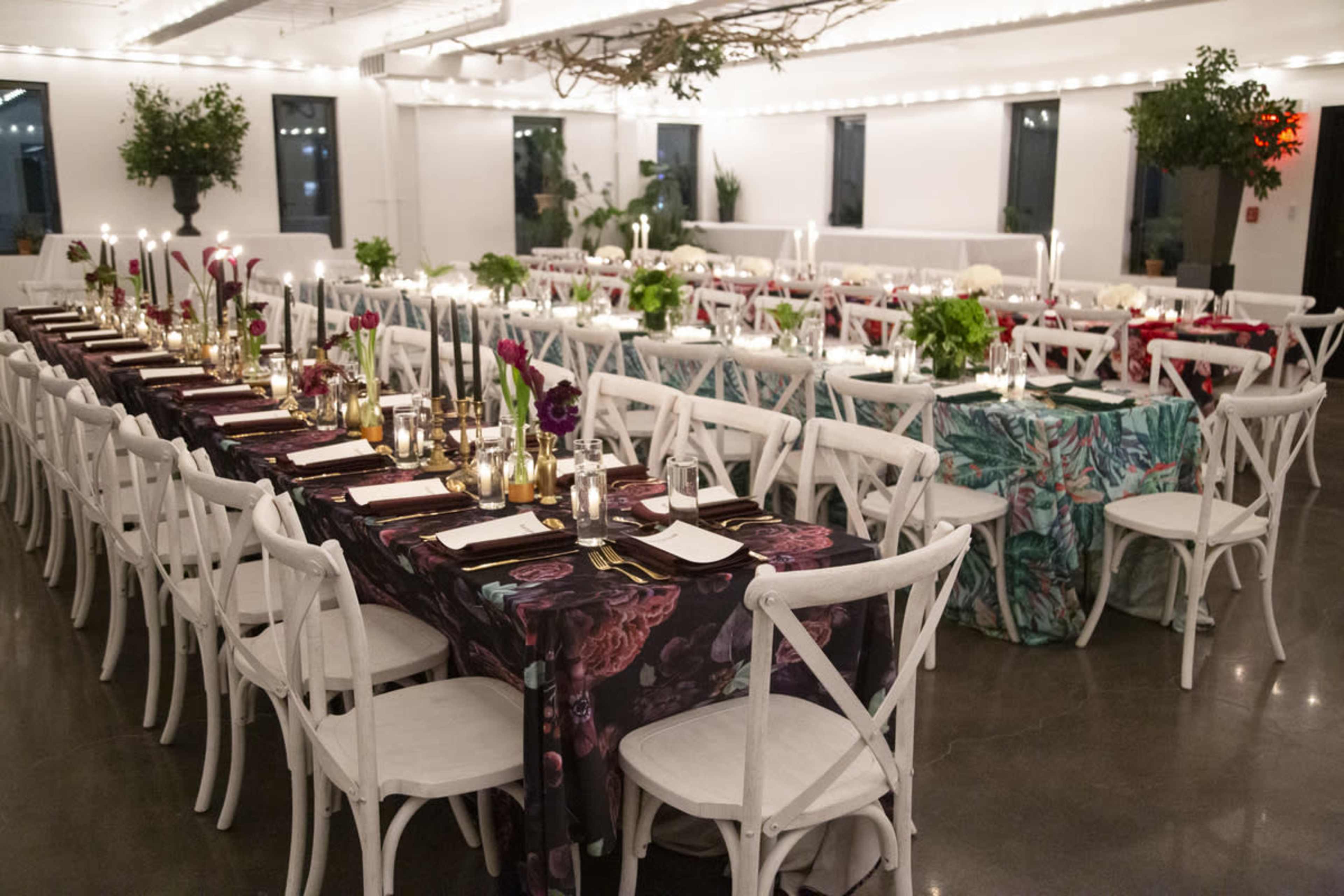 A spacious dining area is set with multiple long tables, each adorned with dark table linens, candles, and centerpieces featuring greenery and flowers.