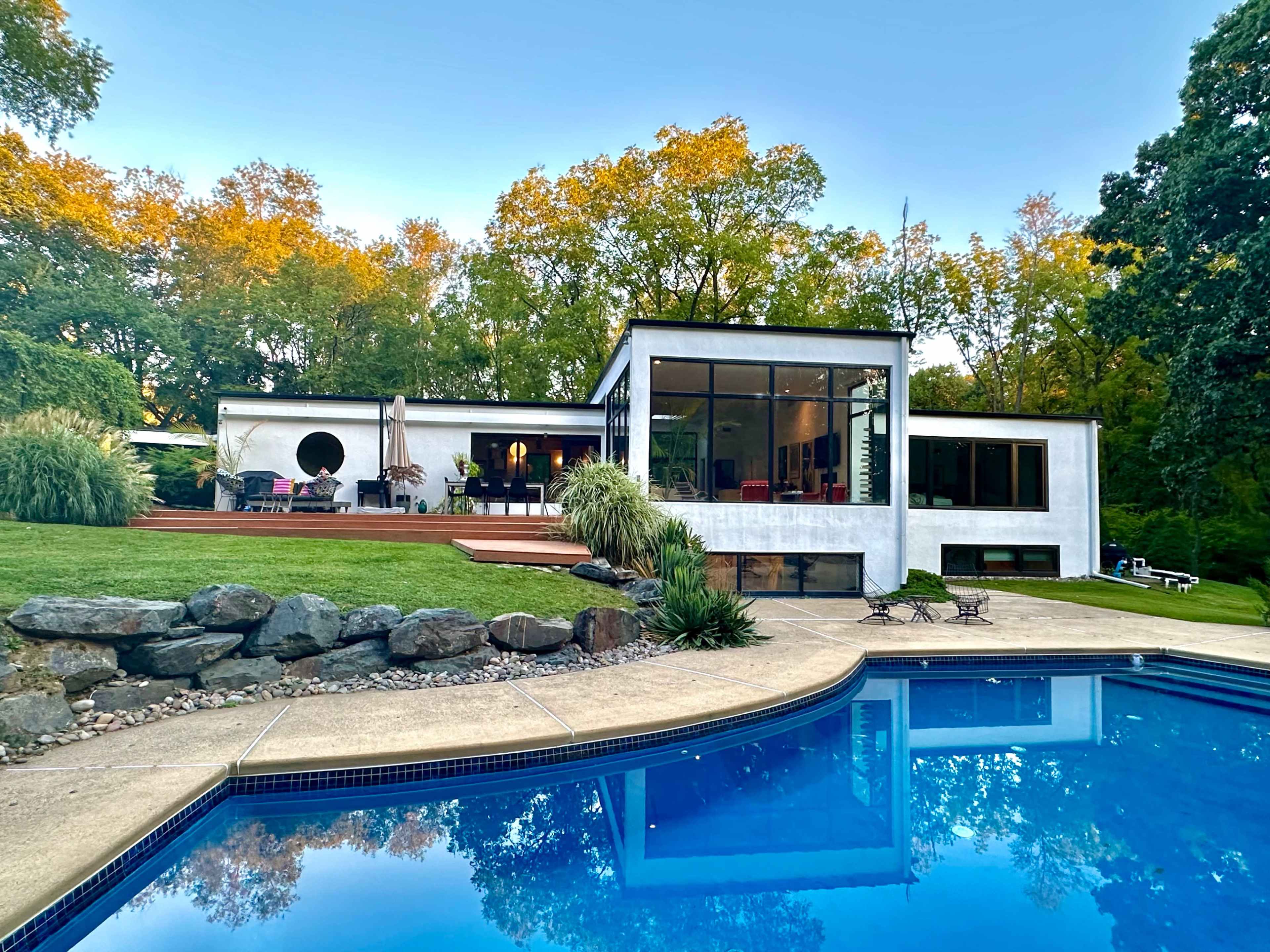 The image depicts a modern white house with large windows, surrounded by greenery and featuring a swimming pool in the foreground.