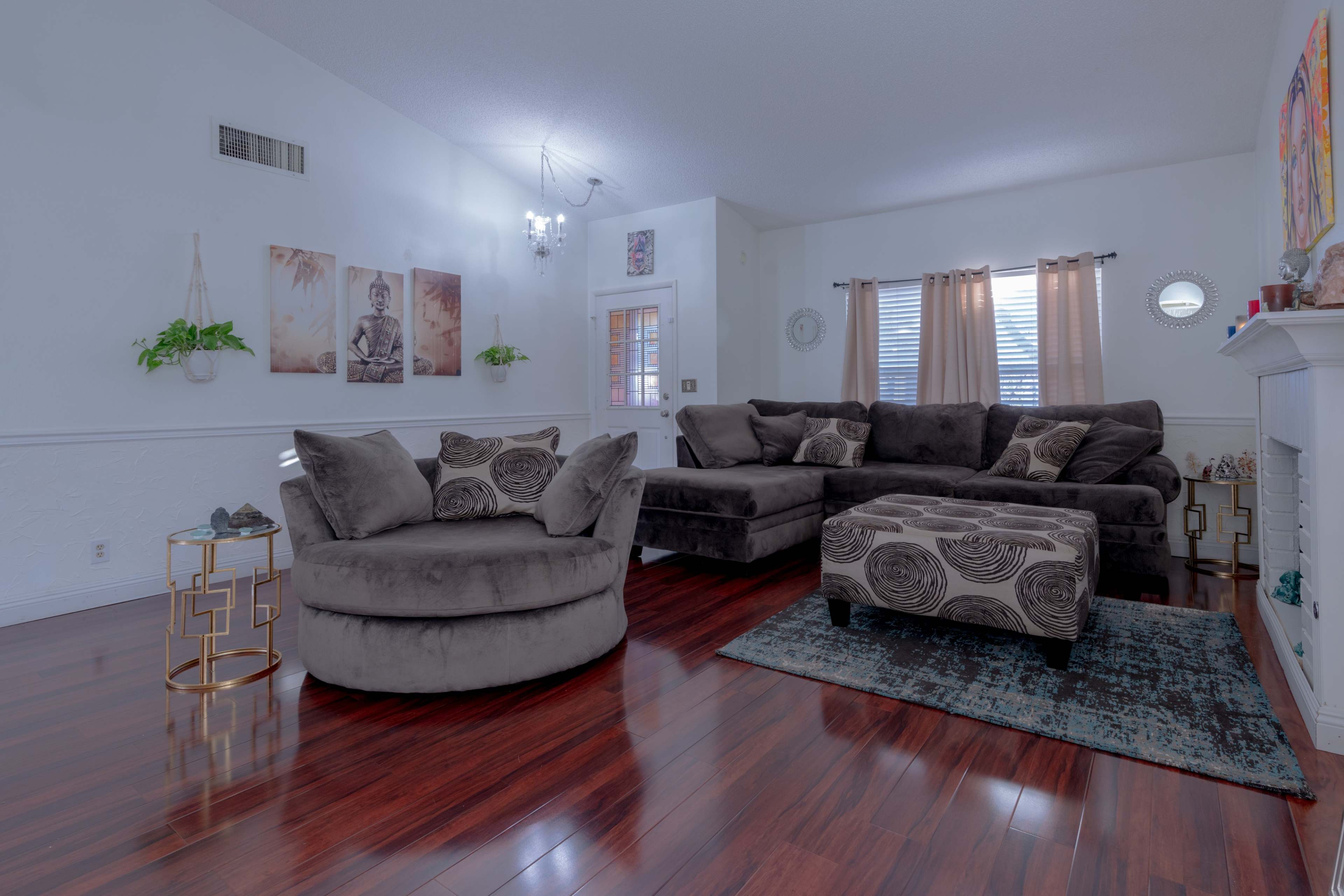The living room features a large sectional sofa, a circular chair, and a coffee table on a hardwood floor, accented by decorative art on the walls and soft natural light coming through the windows.