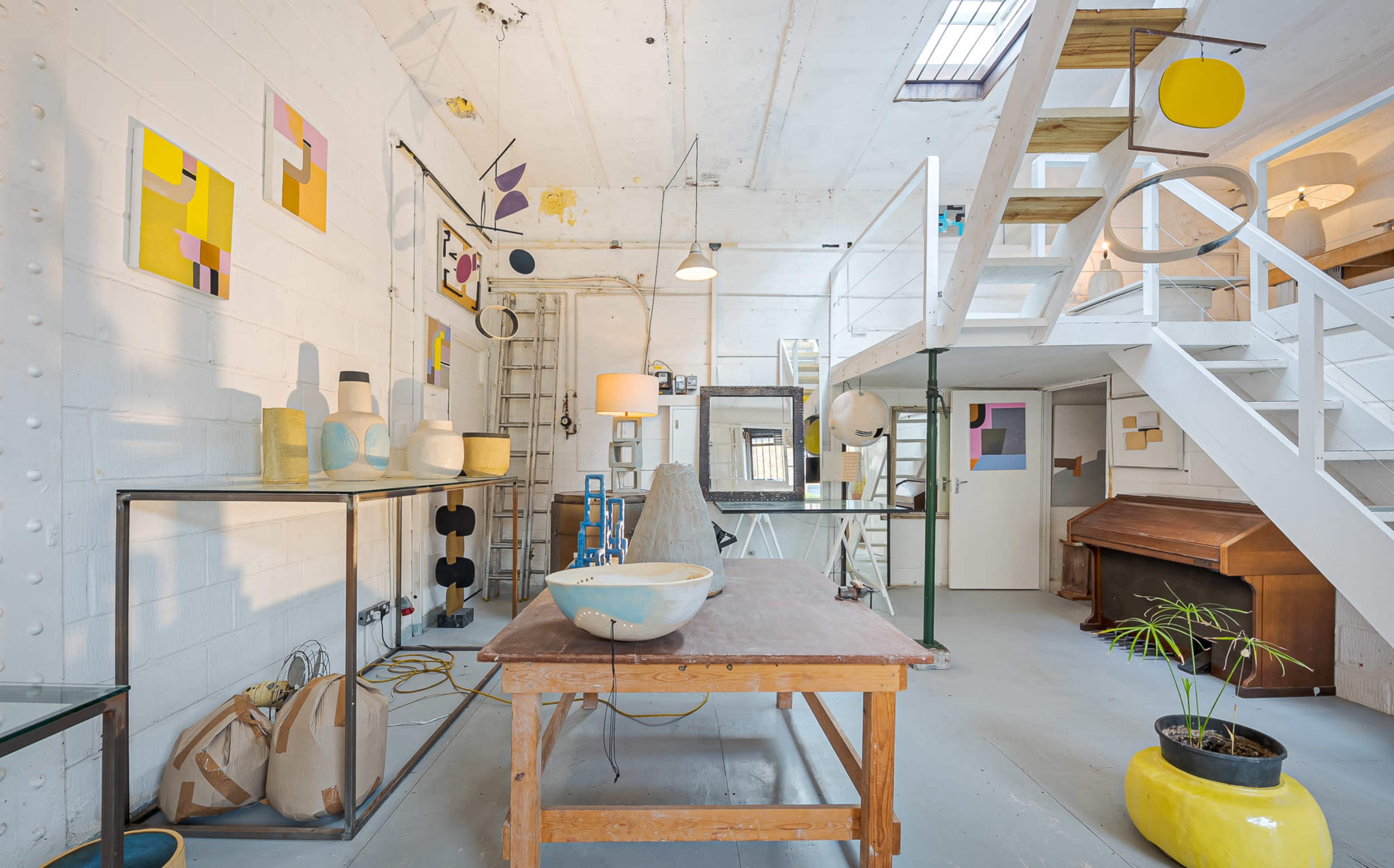 The scene depicts a bright, spacious studio with a wooden table in the foreground, surrounded by various artistic objects, furniture, and artworks on the walls.