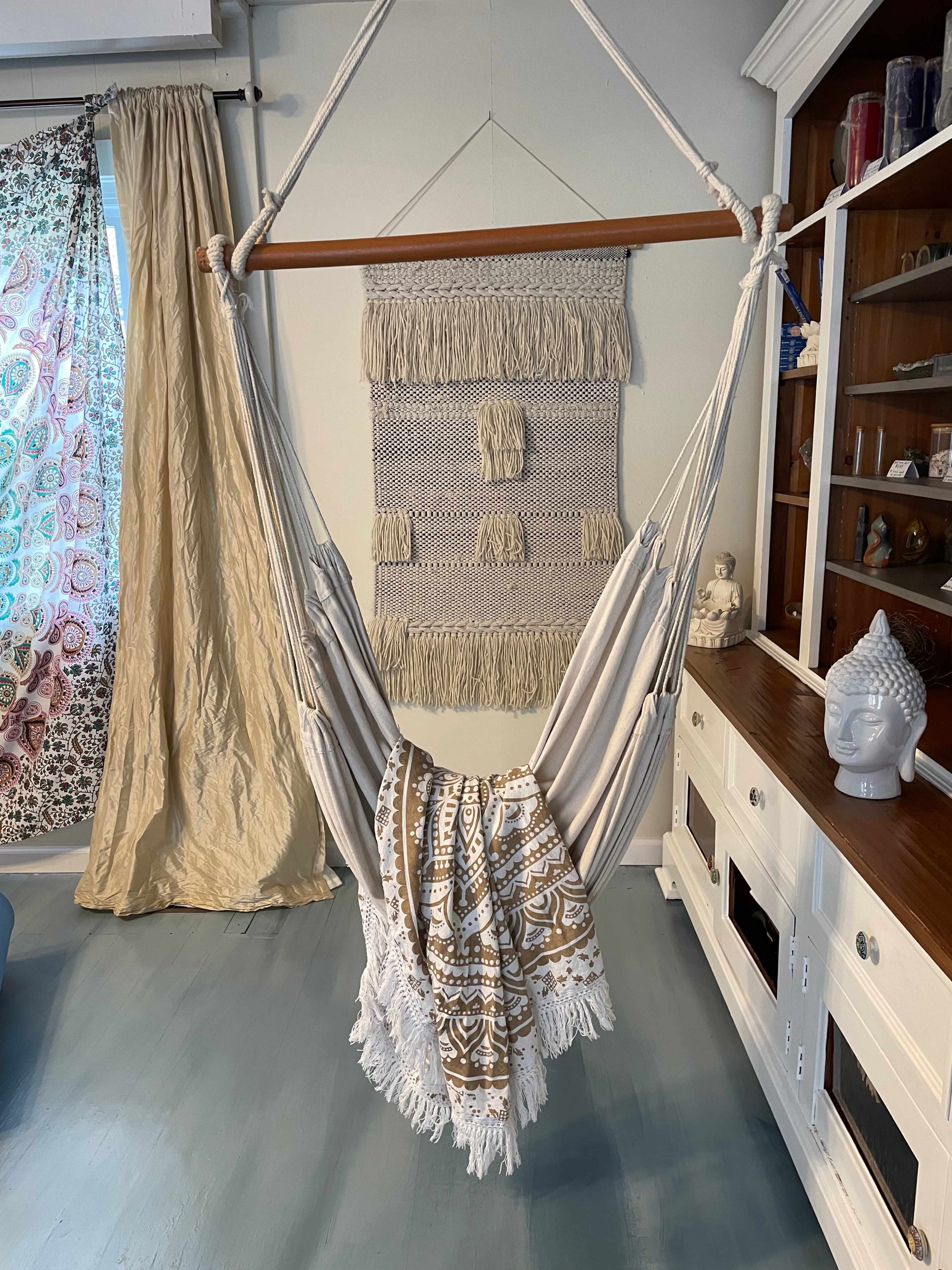 A hammock hangs from a wooden beam, draped with a patterned blanket, in a room featuring decorative wall hangings and light-colored furniture.