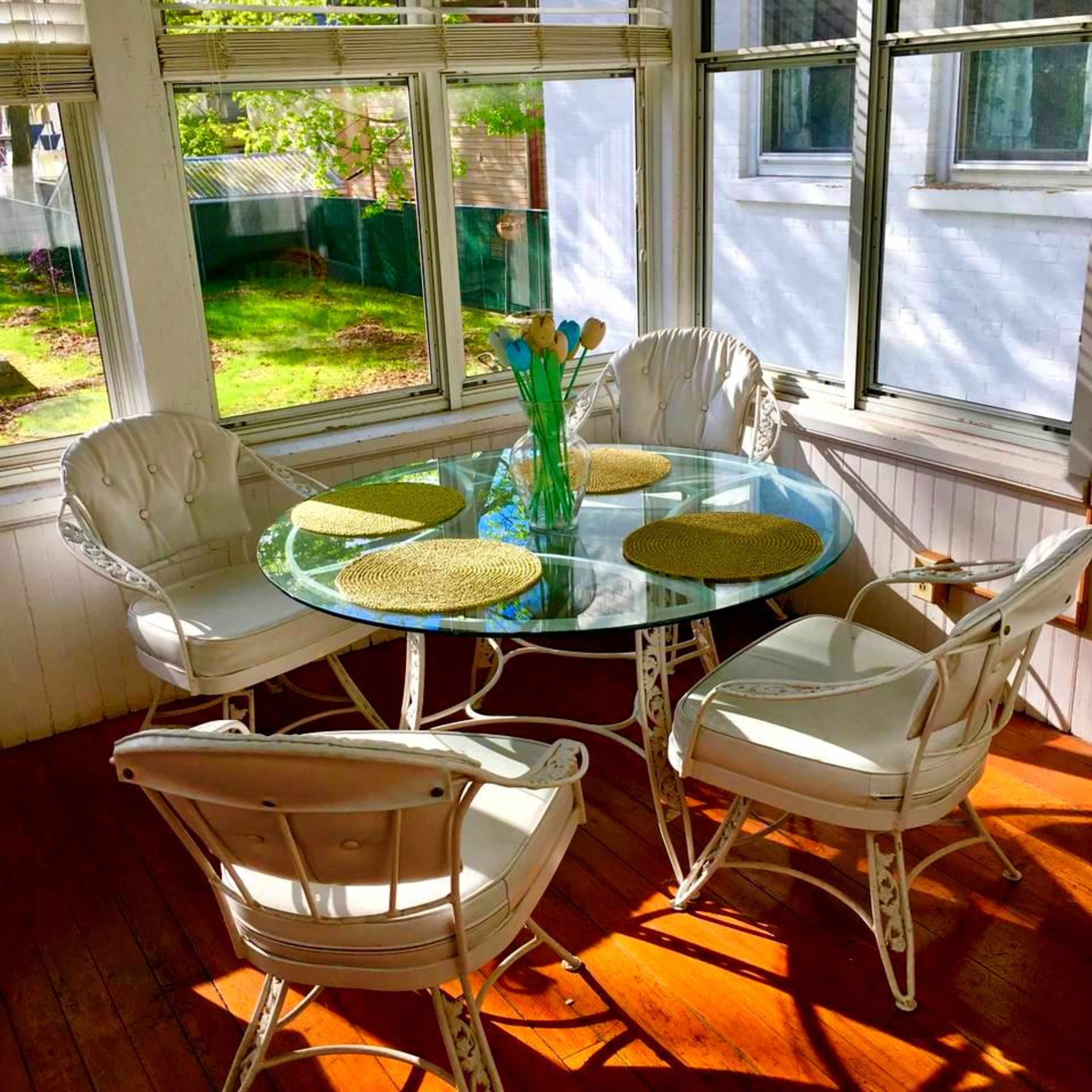 A glass-top dining table surrounded by four white metal chairs is set in a sunlit corner surrounded by windows.