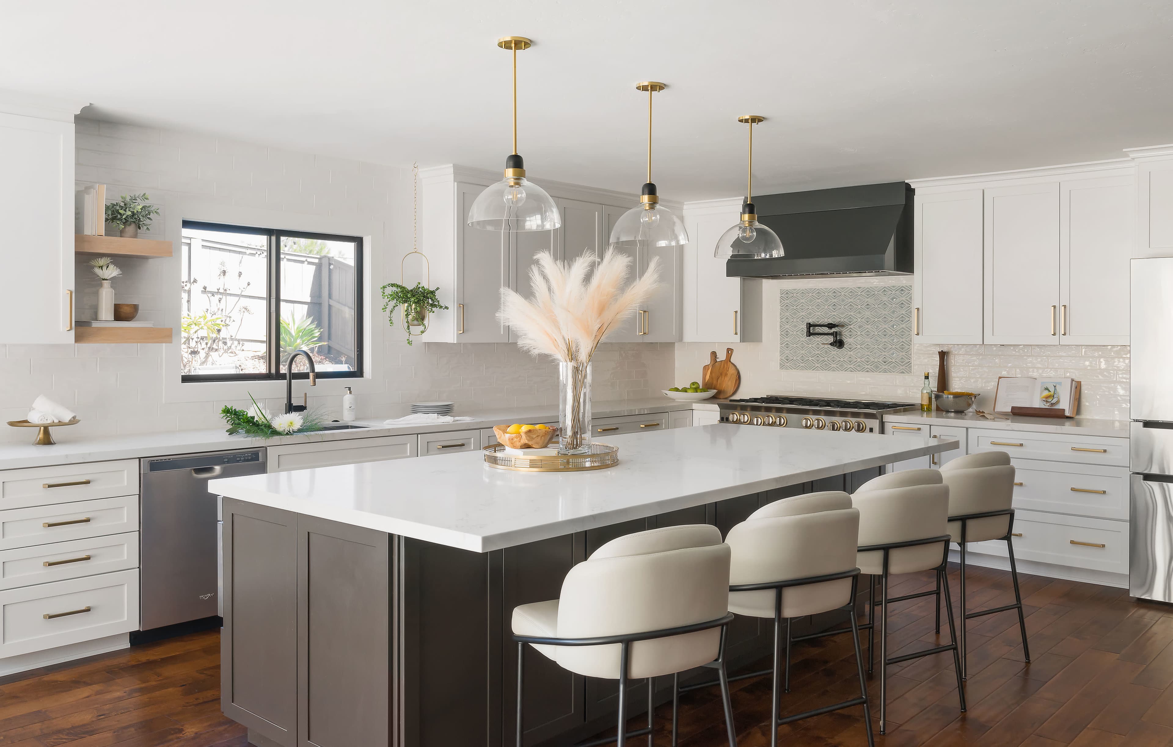 A modern kitchen features a large island with seating, white cabinets, and gold accents, illuminated by pendant lights.