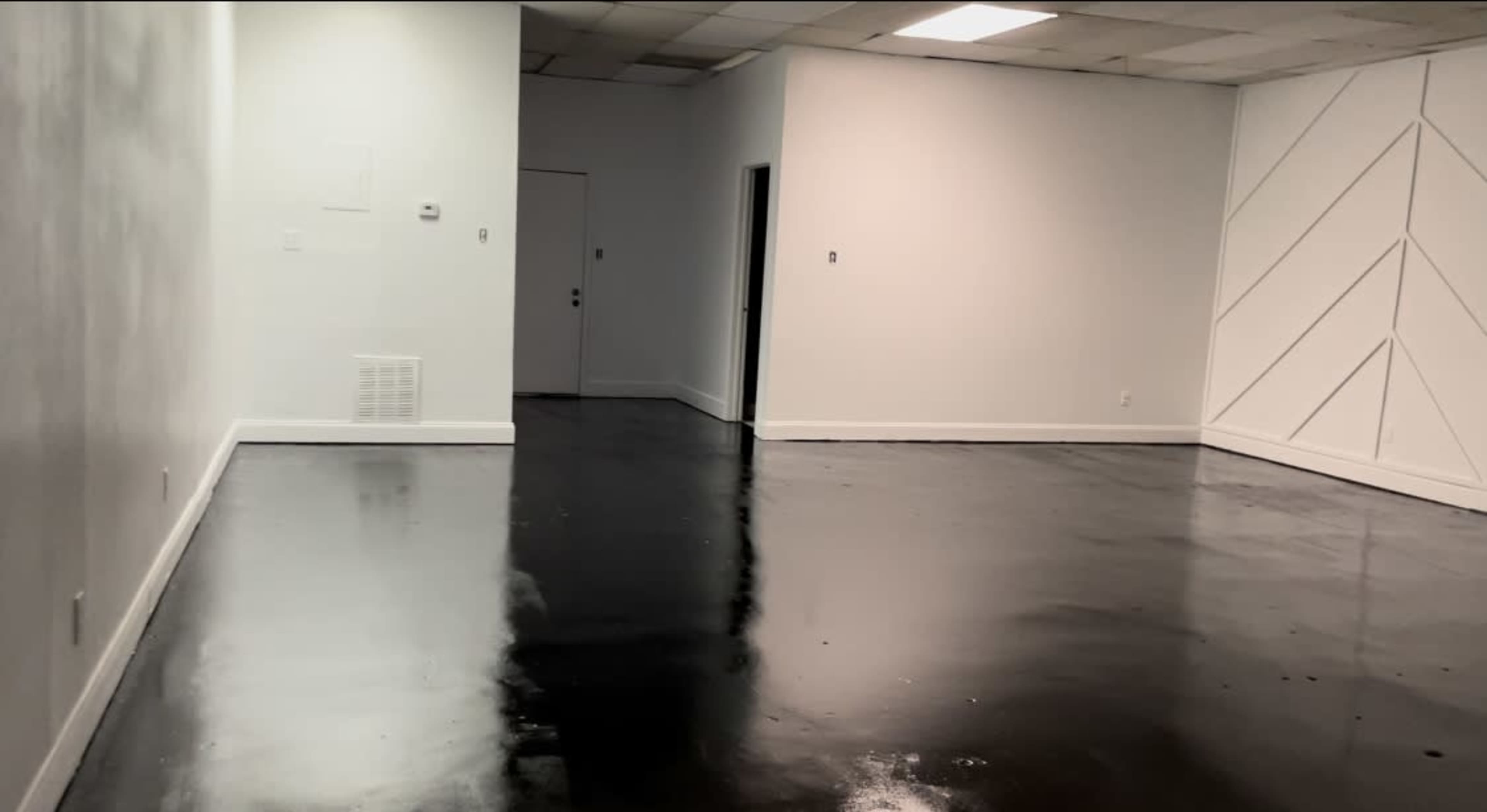 The image shows a sparsely furnished room with white walls and a glossy black floor, featuring a door on the left and a geometric pattern on the right wall.