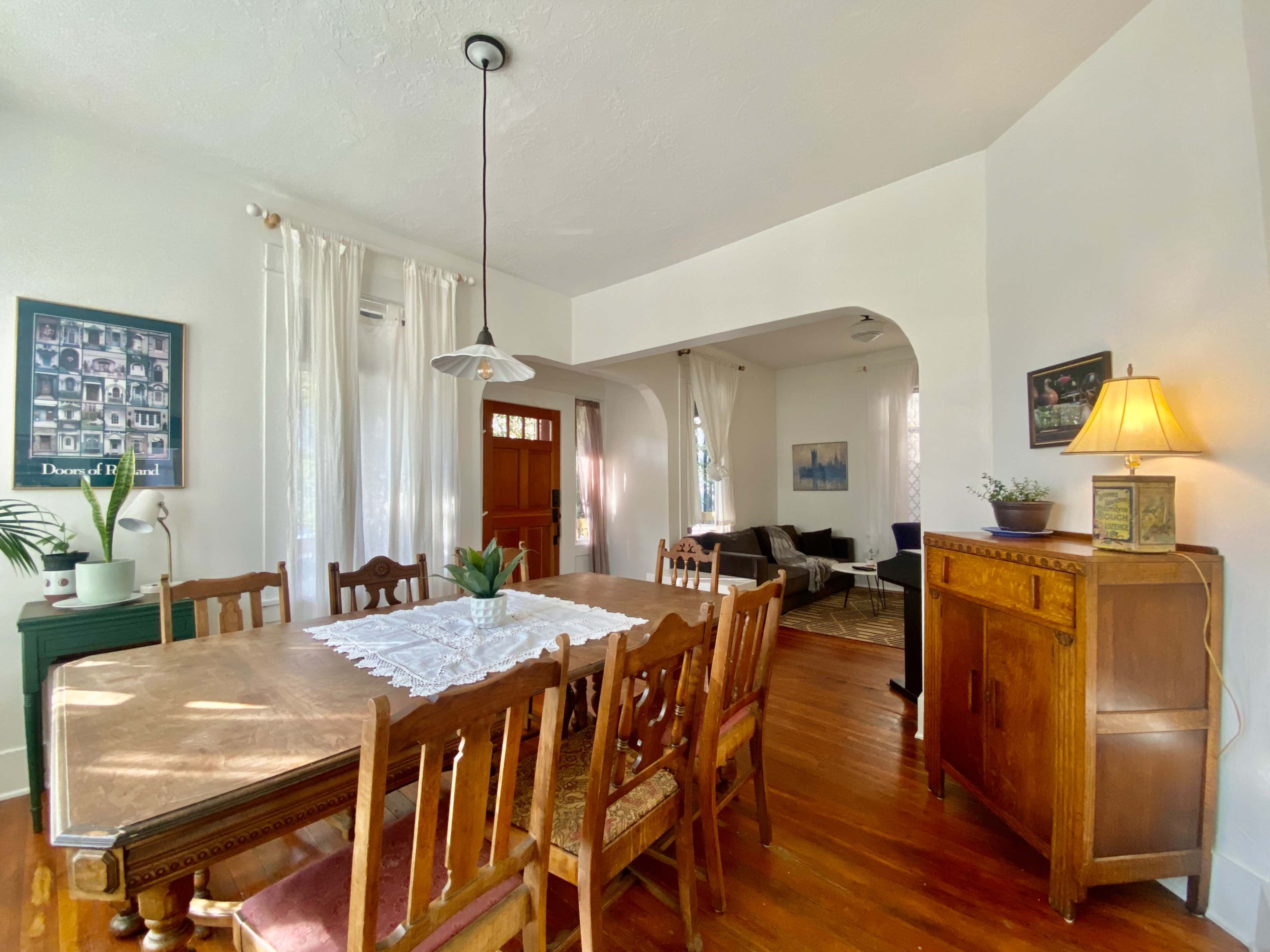 A wooden dining table surrounded by chairs is set in a well-lit room with a sideboard, a lamp, and large windows dressed with sheer curtains.