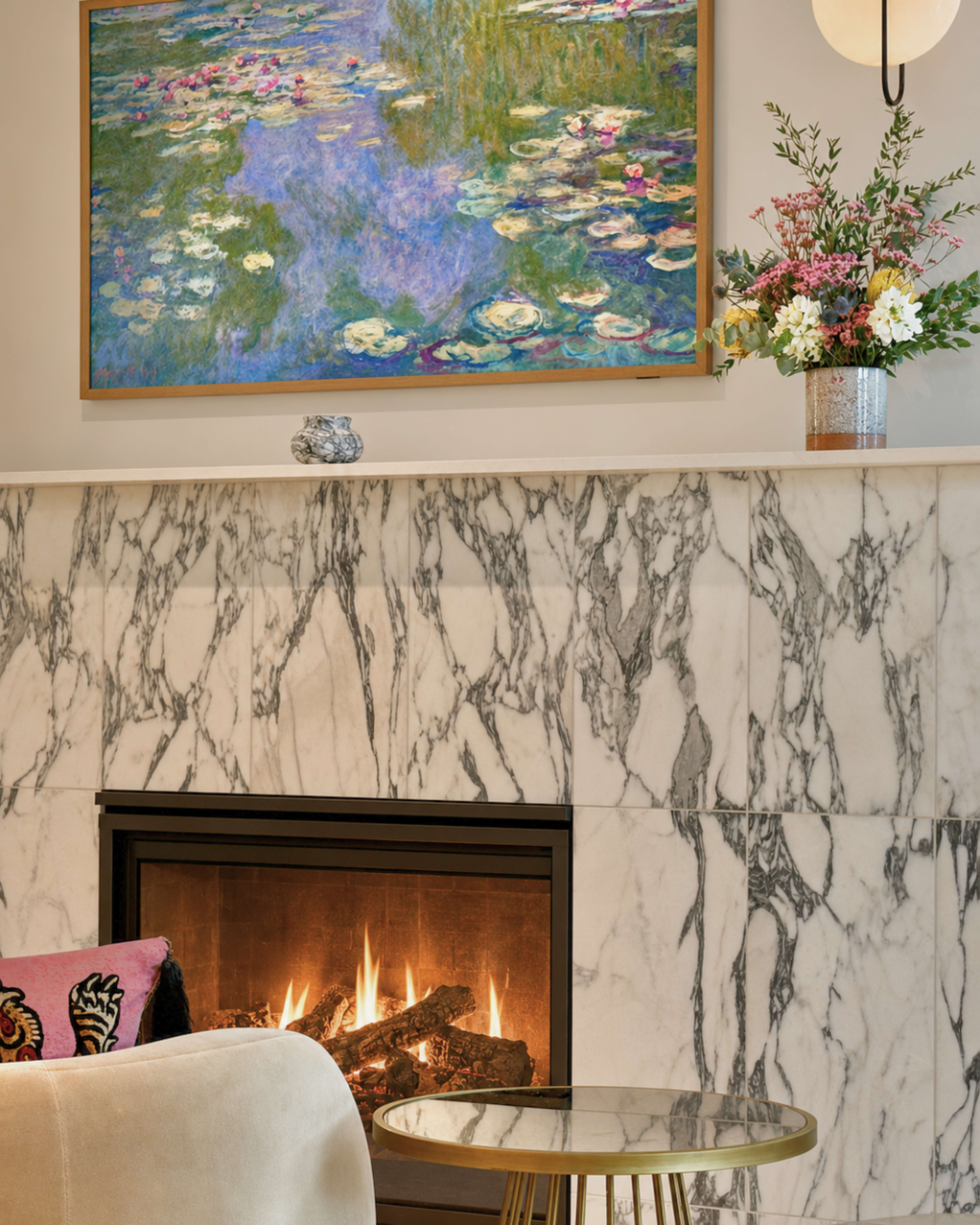 The image shows a modern living room featuring a marble fireplace with a lit fire, a colorful painting of water lilies hanging above it, and a decorative table with flowers.