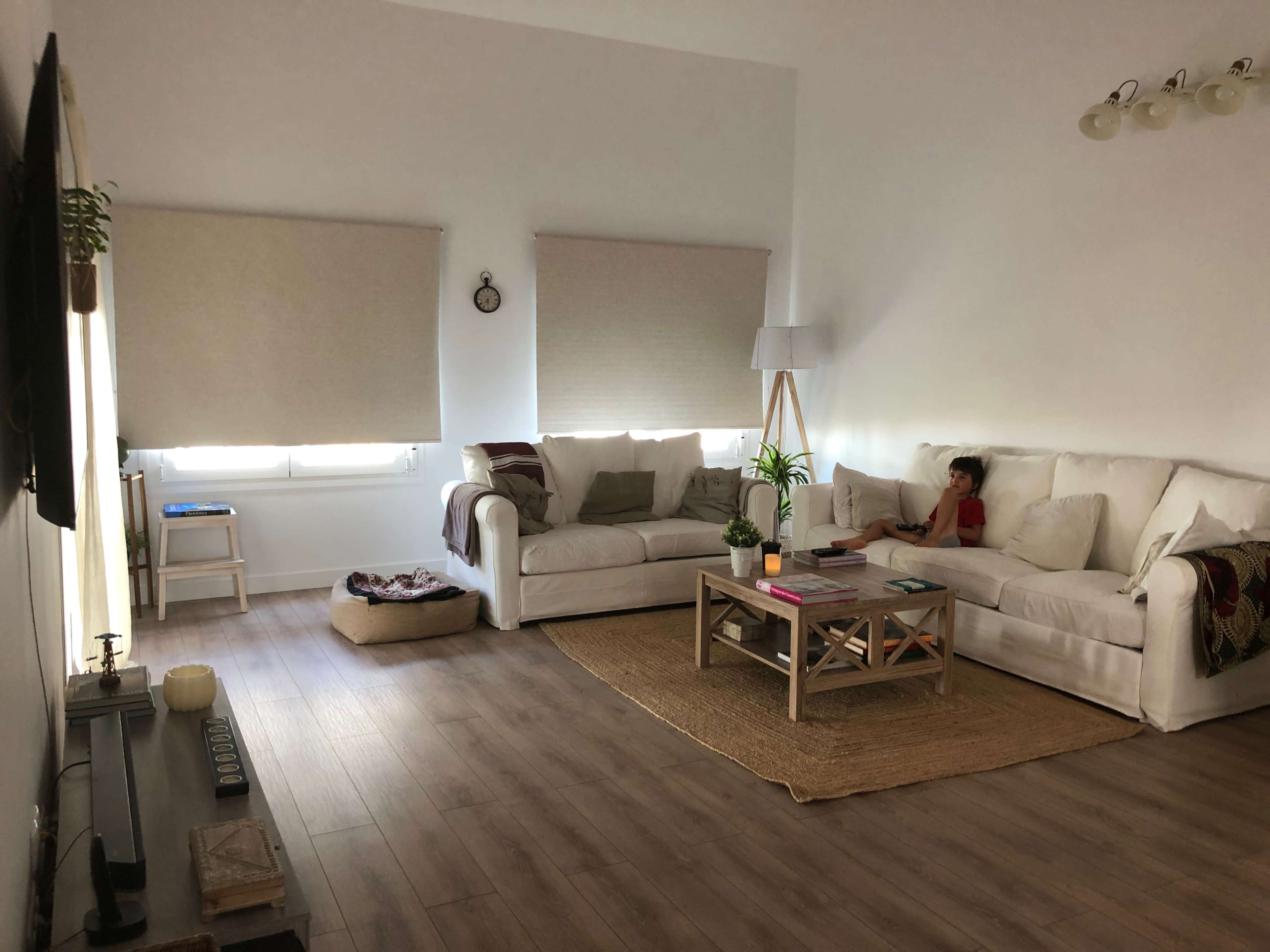 A child sits on a white sectional sofa in a well-lit living room with natural wood flooring and minimal decor.