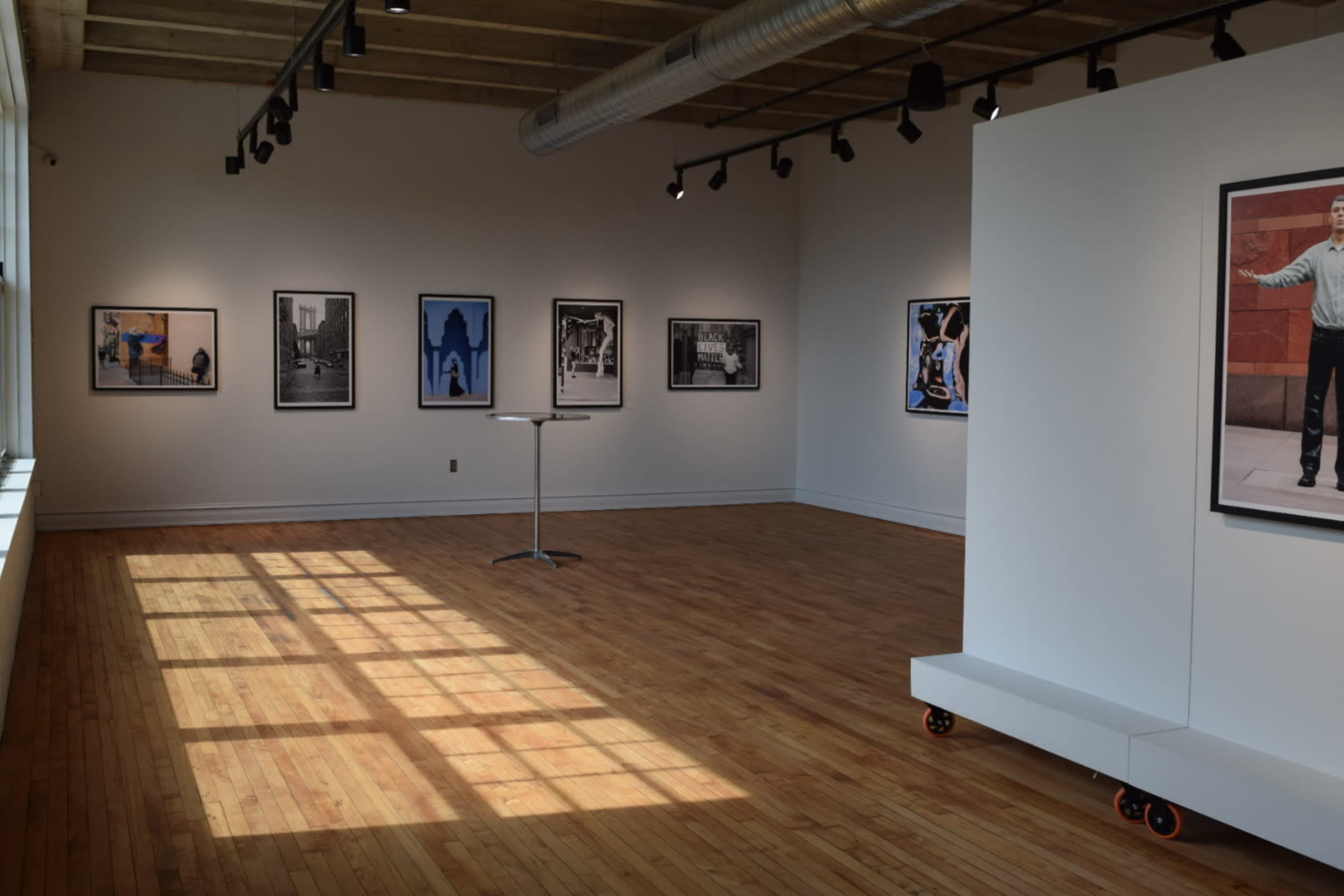 The image shows an art gallery with several black-and-white photographs displayed on the walls and a single table in the center, illuminated by natural light from large windows.