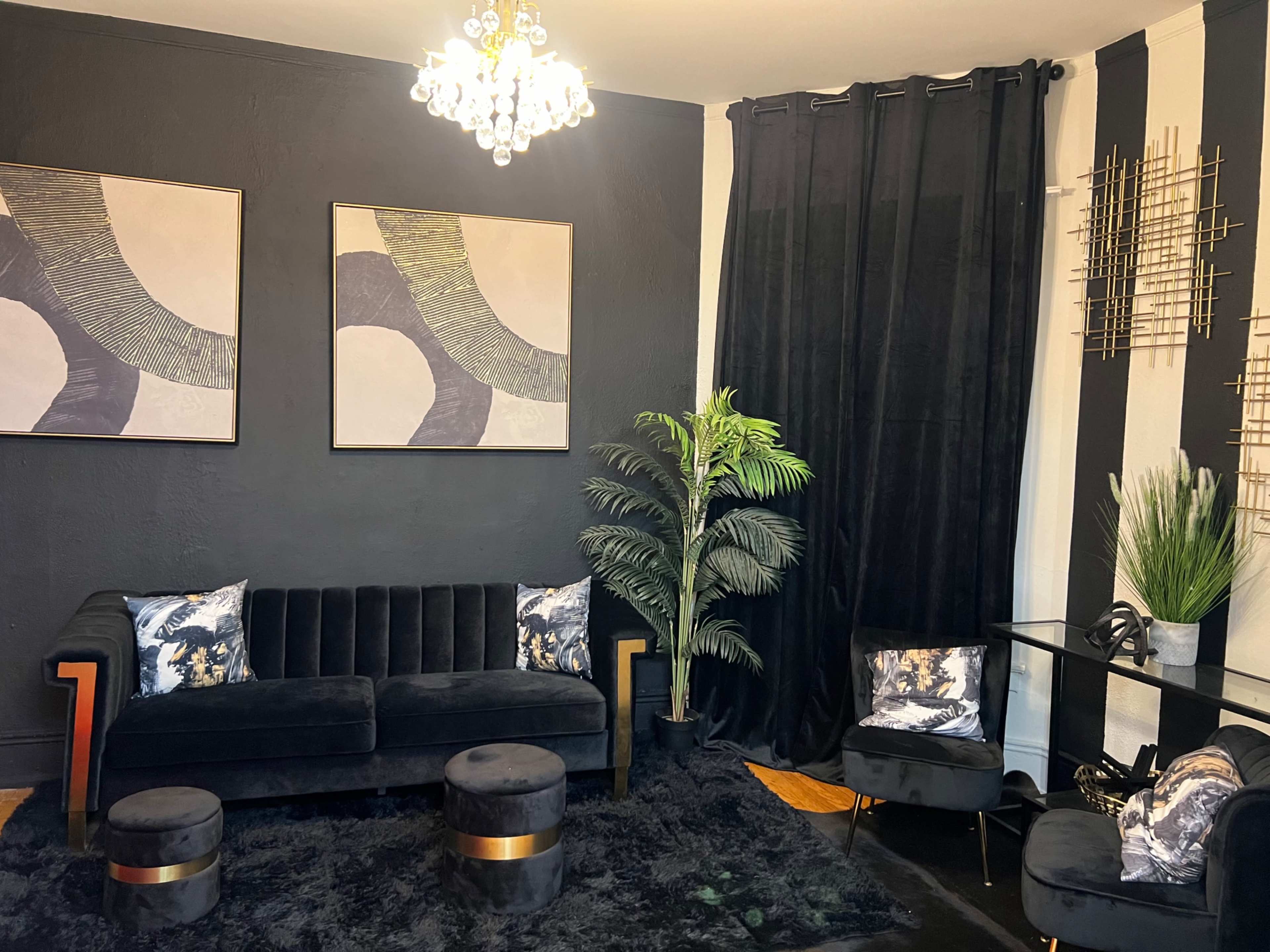 The image shows a modern living room with a black velvet sofa, decorative pillows, and a large plant, complemented by abstract wall art and a chic chandelier.