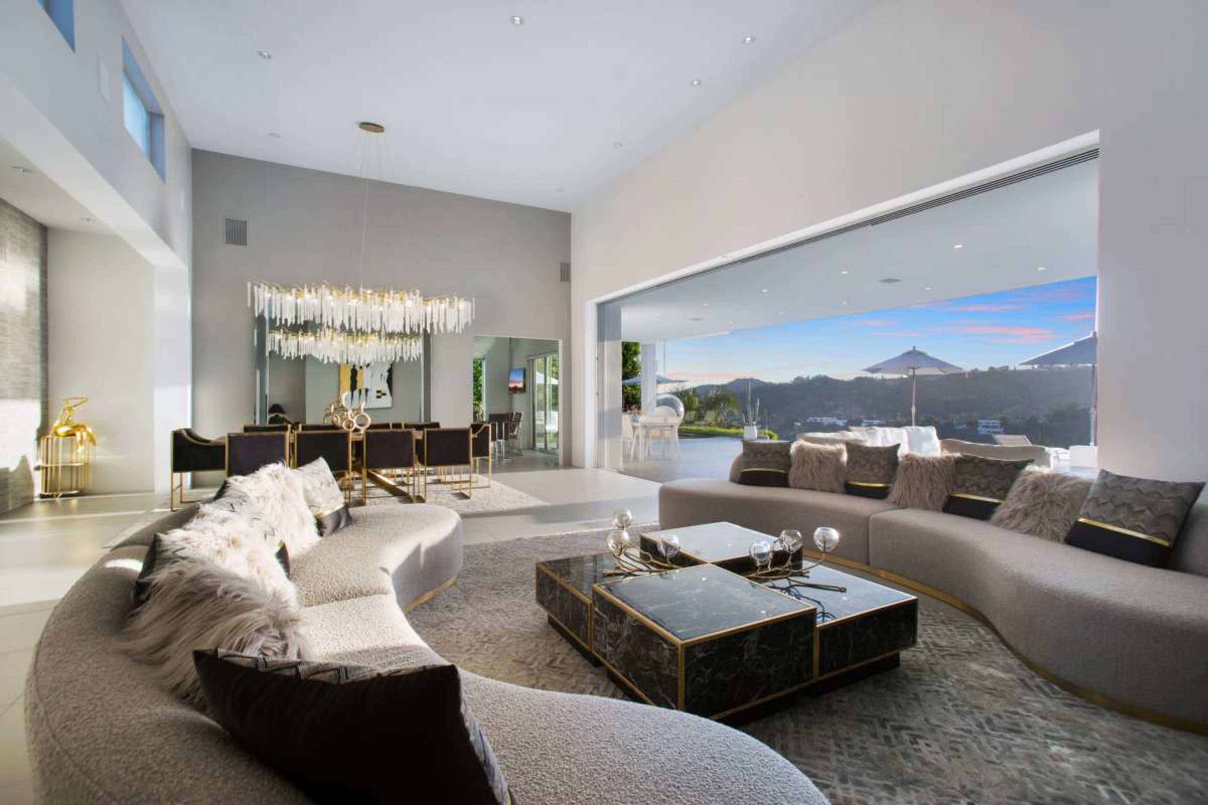 The image shows a modern living room with a circular sectional sofa, a coffee table, and a dining area featuring a chandelier and large windows overlooking an outdoor space.