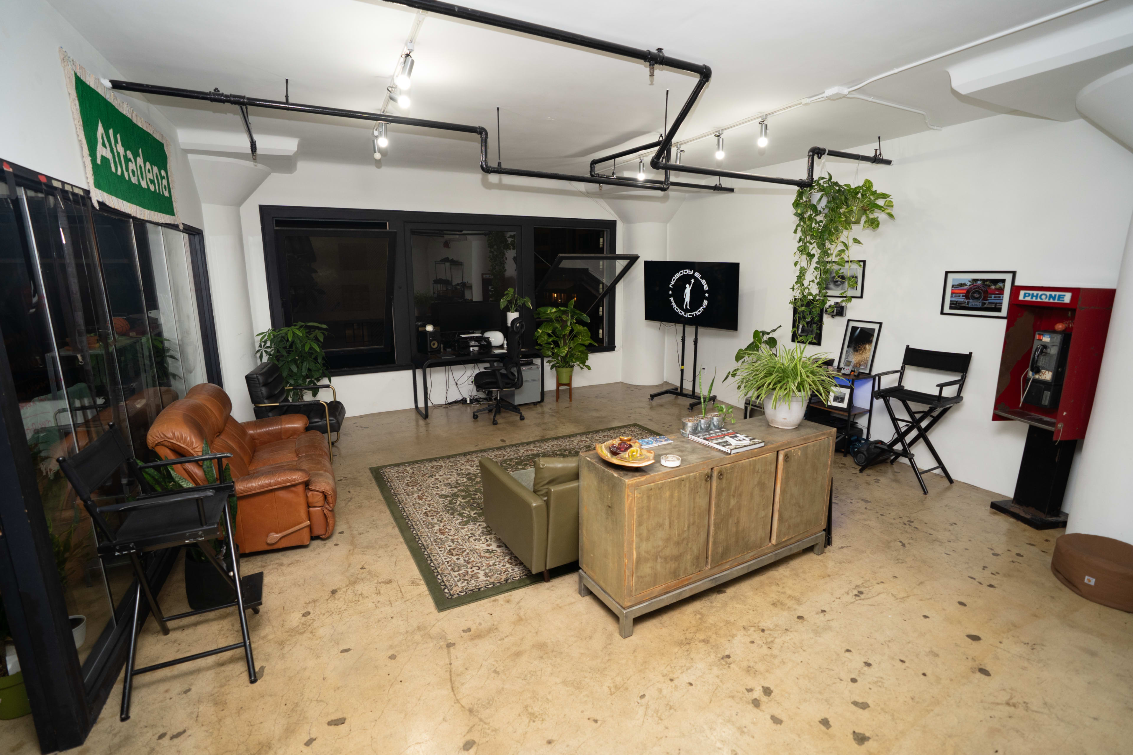 The image shows a modern office space featuring seating areas, a large window, a desk with a chair, and decorative plants.