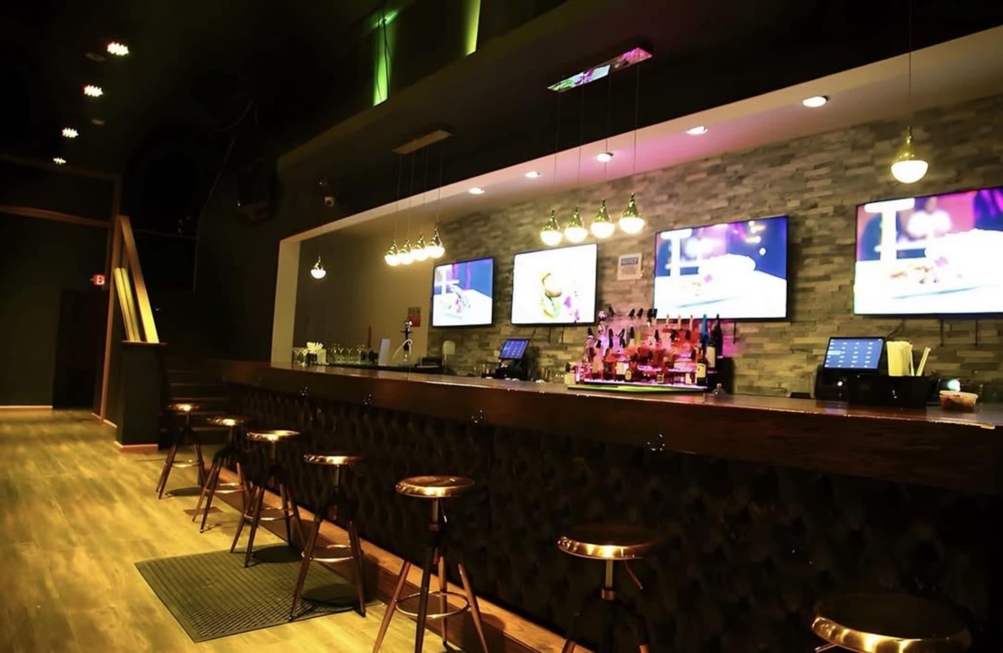 The image shows a bar with wooden flooring, a long wooden counter, stools, and several television screens displaying menus in a dimly lit setting.