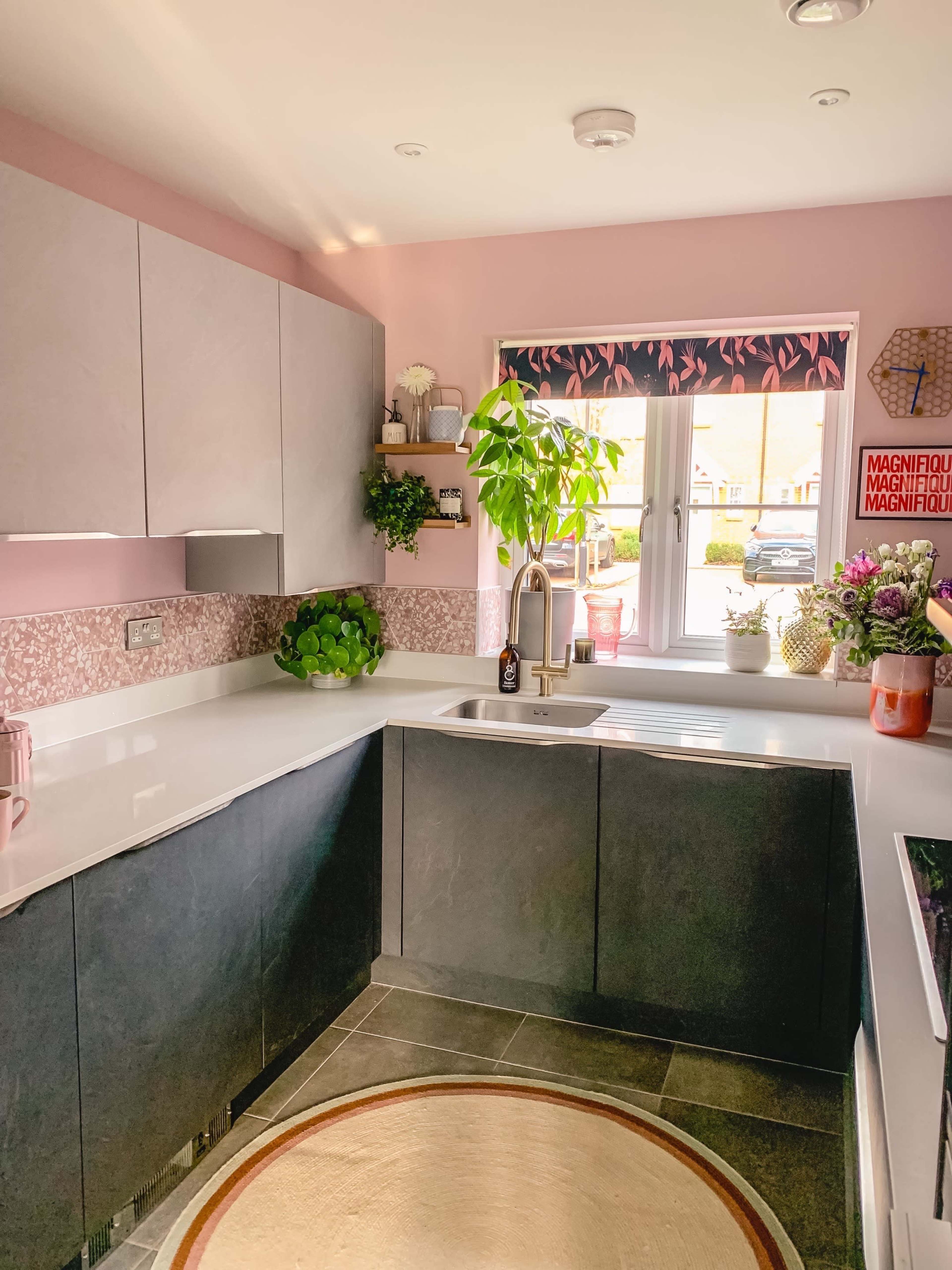 A modern kitchen features light pink walls, gray cabinets, and a window with plants on the sill.