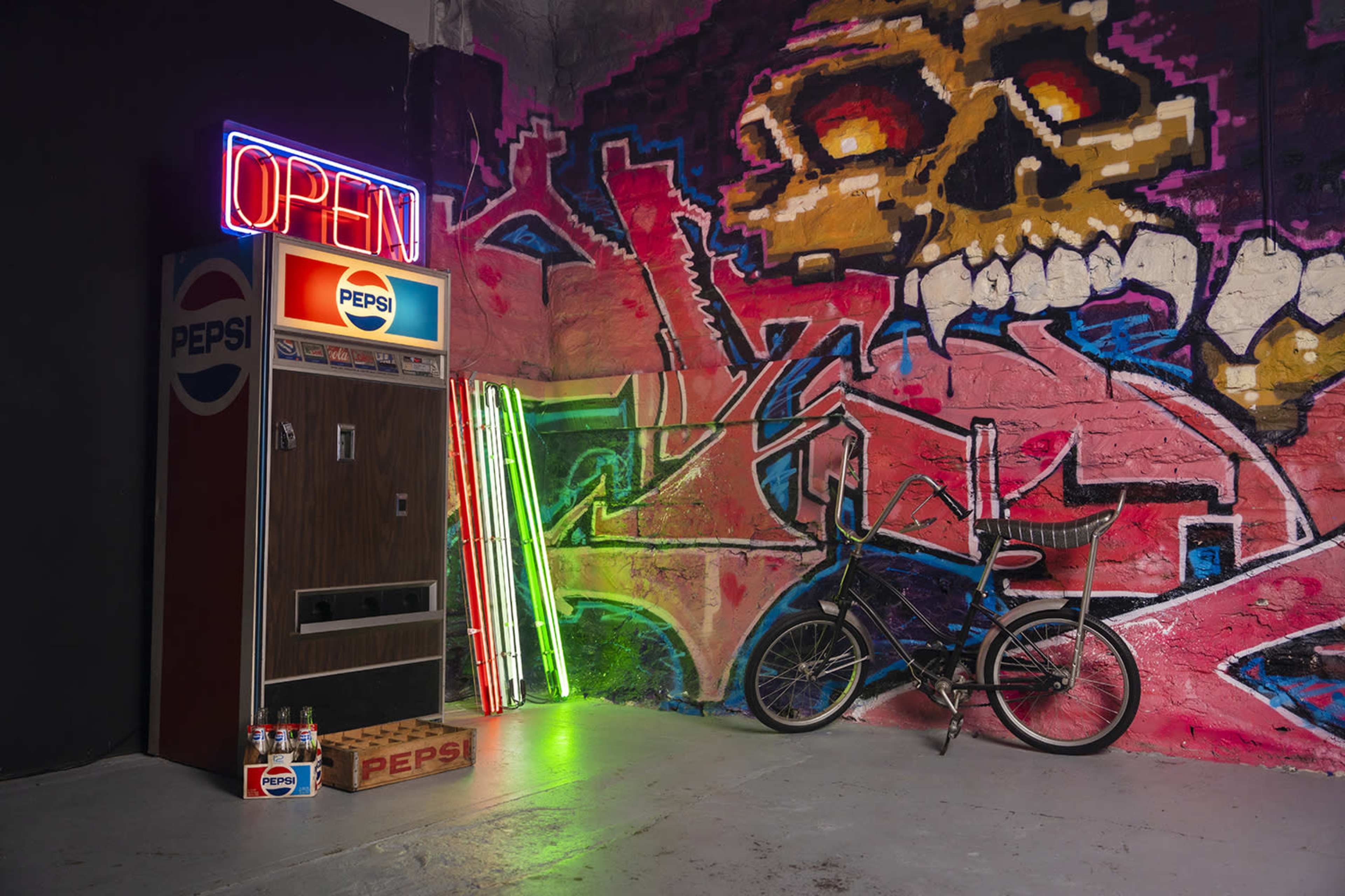 A Pepsi vending machine beside a bicycle and illuminated neon strips against a graffiti-covered wall featuring a skull design.