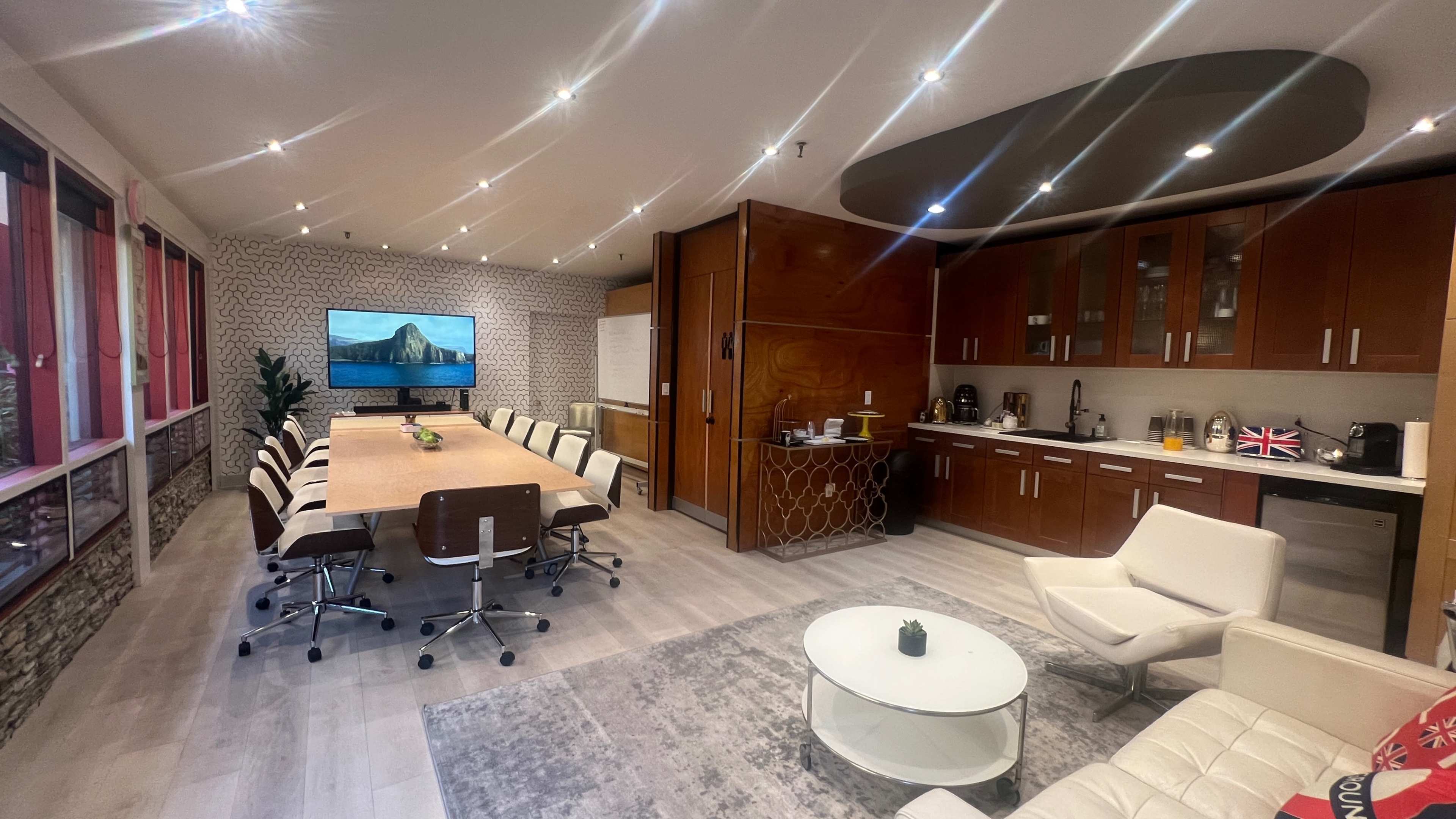 A modern conference room with a large table surrounded by chairs, a kitchenette, and a television displaying a scenic landscape.
