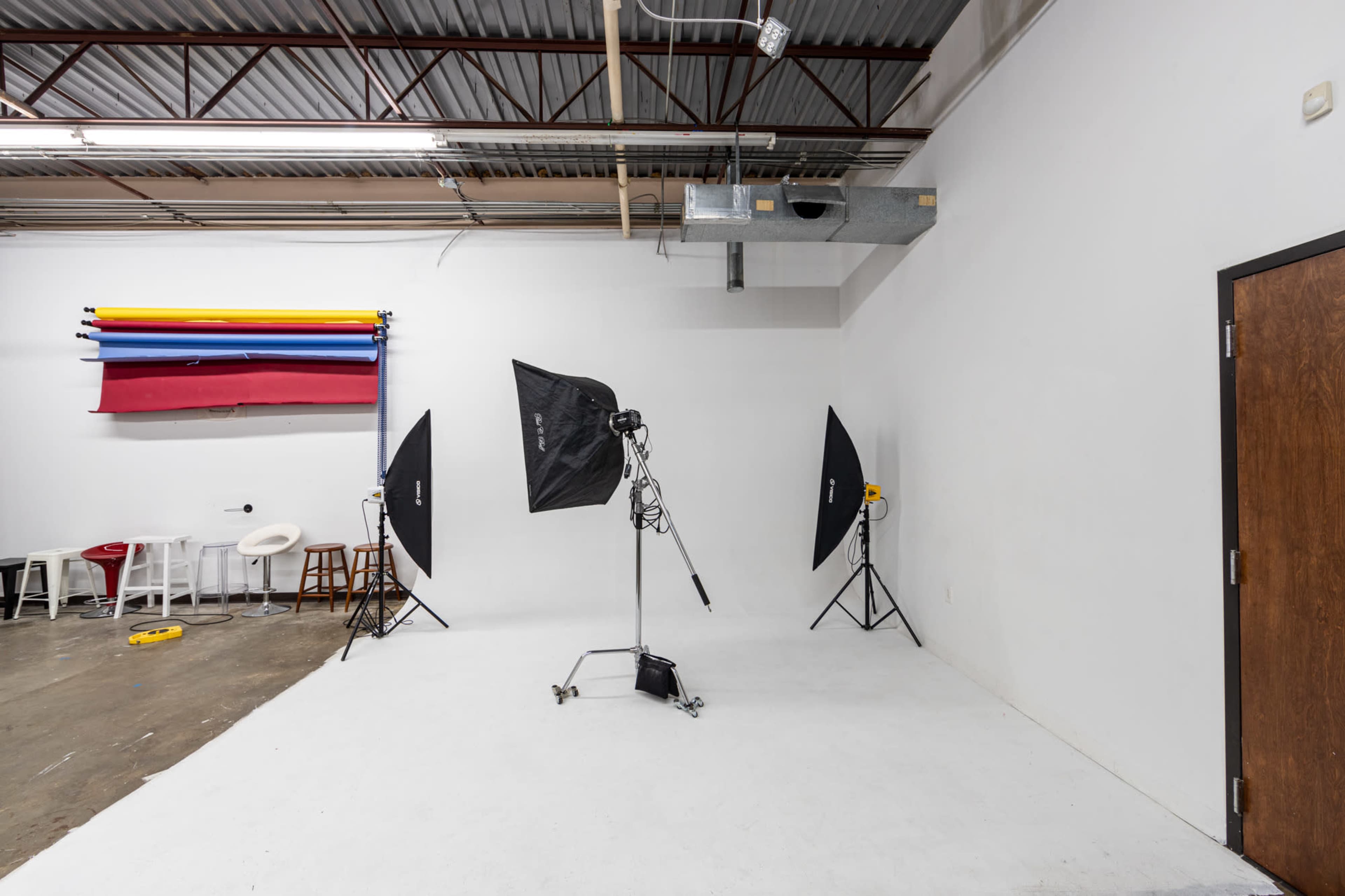The image shows a photography studio with three light setups, a backdrop of colored rolls, and empty furniture against a white wall.