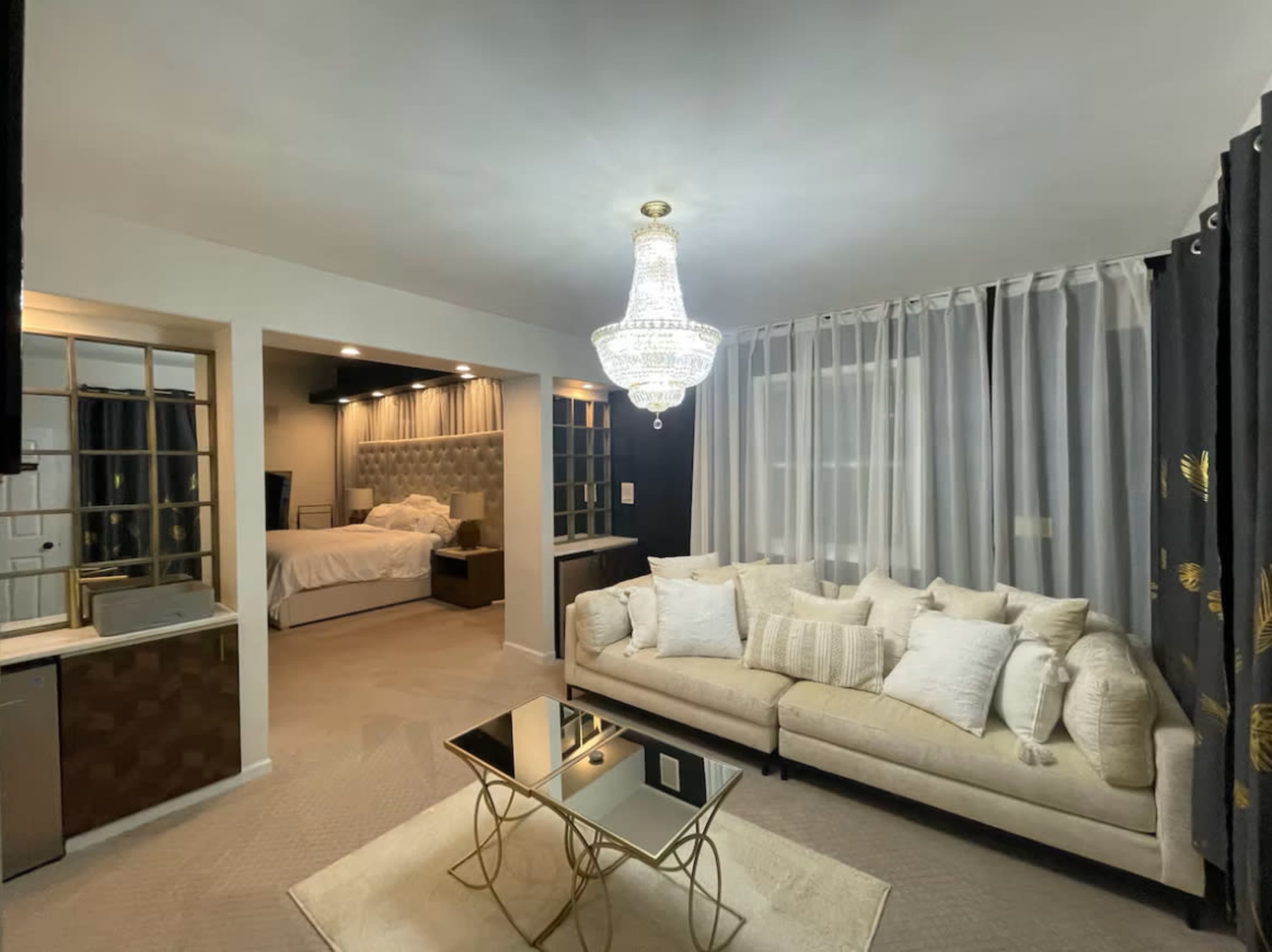 The image shows a well-lit room featuring a modern sofa with decorative pillows, a glass coffee table, and a view of a bedroom area in the background.
