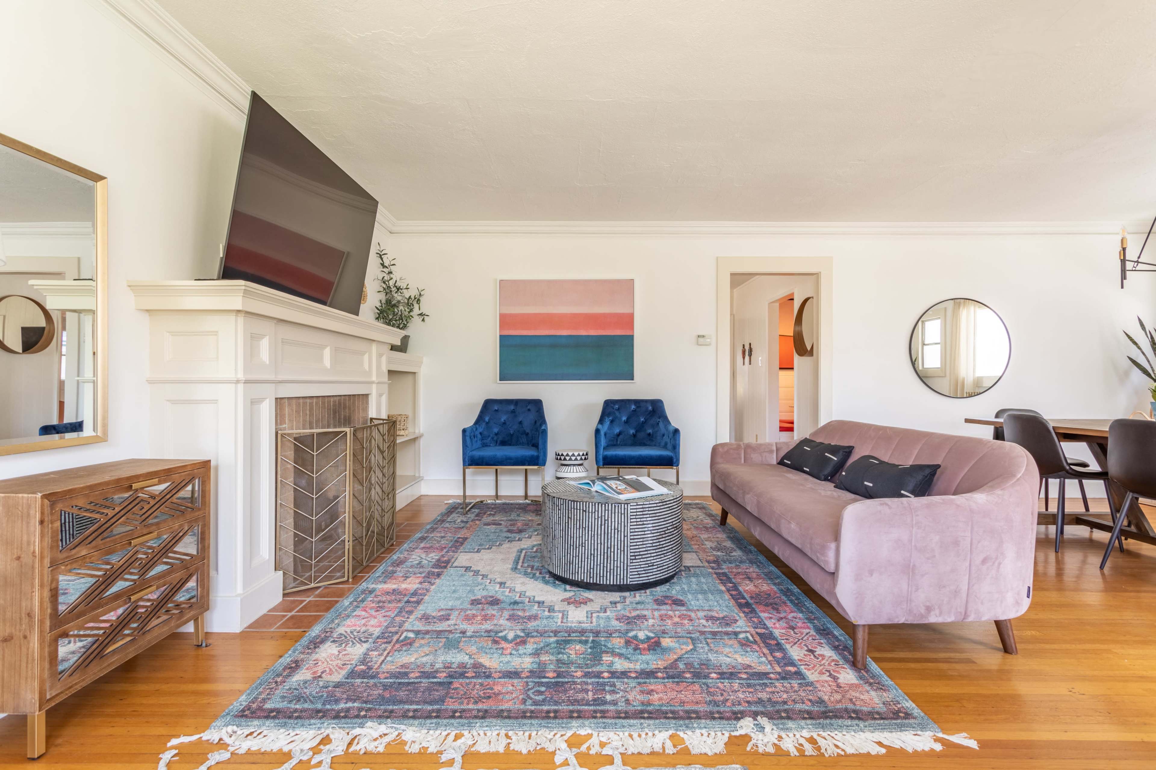 A modern living room features a pastel artwork above a pink sofa, flanked by blue armchairs and a wooden side table on a patterned rug.