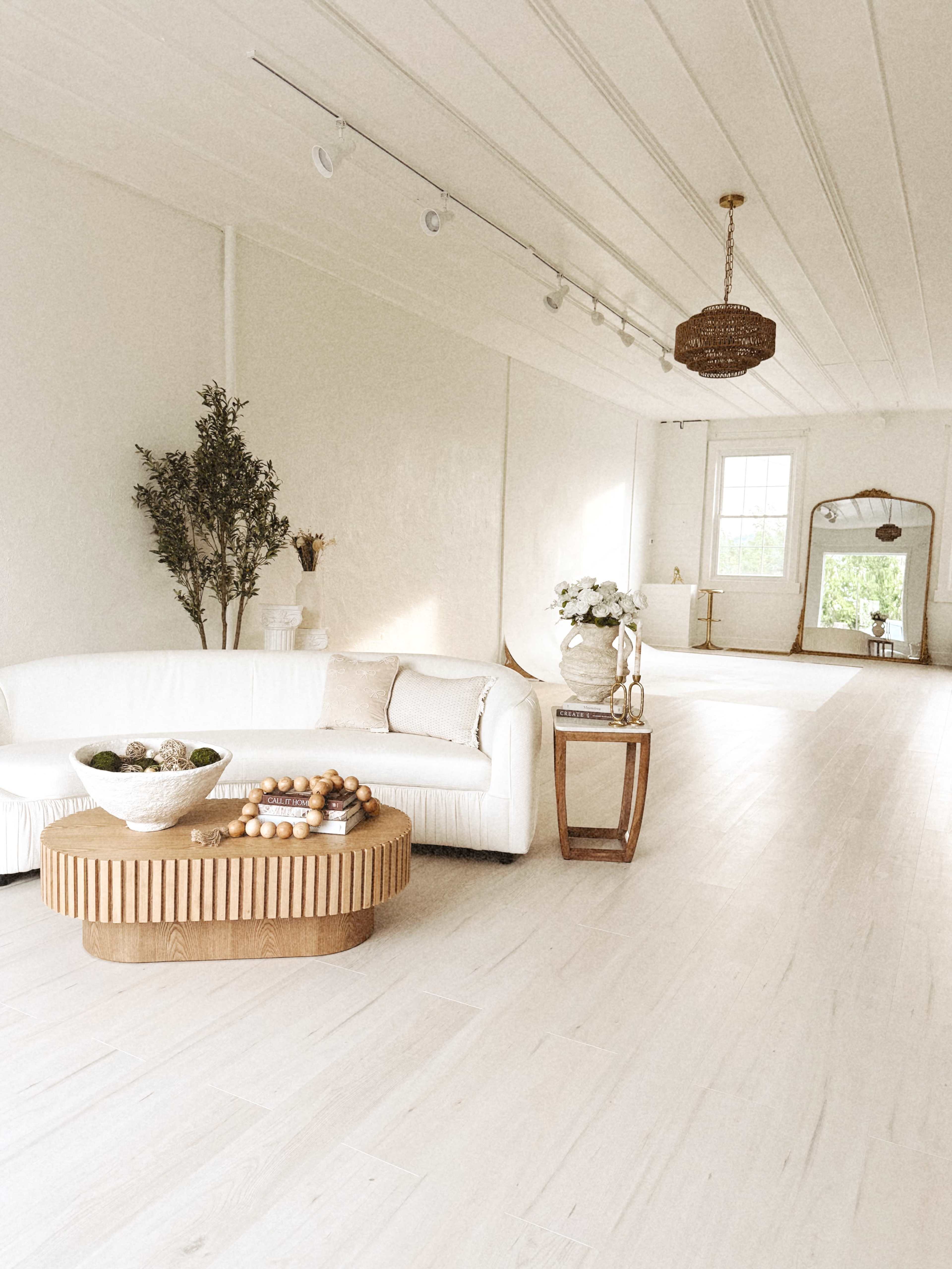 A bright, spacious room features a white curved sofa, a wooden coffee table adorned with decorative items, and large windows providing natural light.