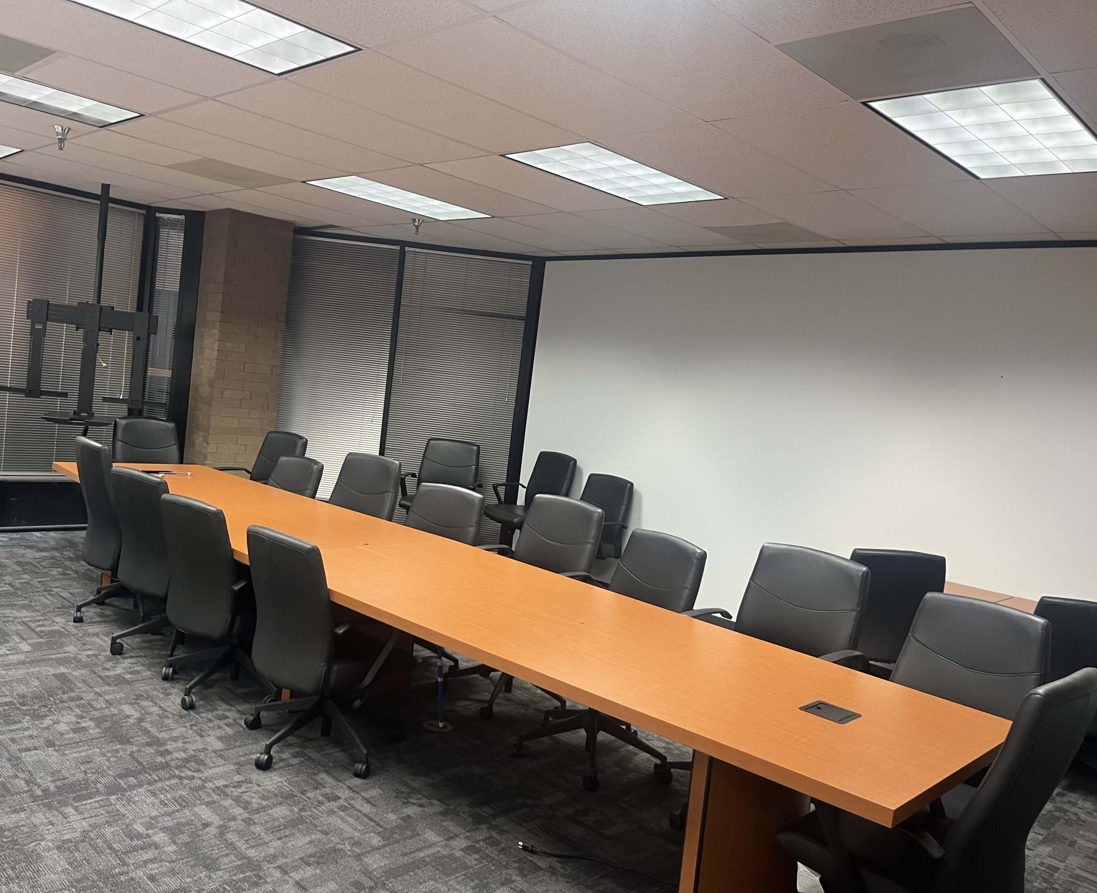 The image shows a conference room with a long wooden table surrounded by black office chairs and a blank white projection wall.