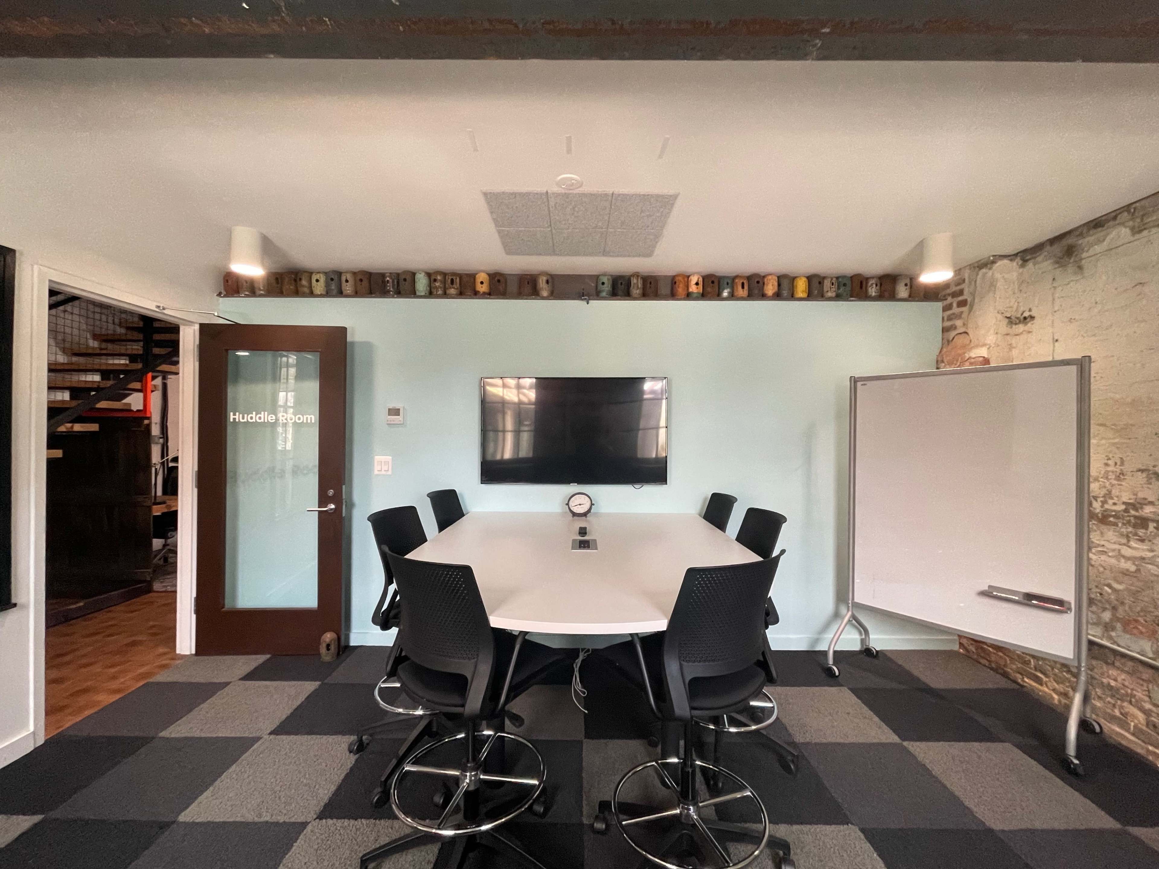 A huddle room features a circular table surrounded by high-back chairs, a wall-mounted television, a whiteboard, and a clock, with exposed brick and a staircase in the background.