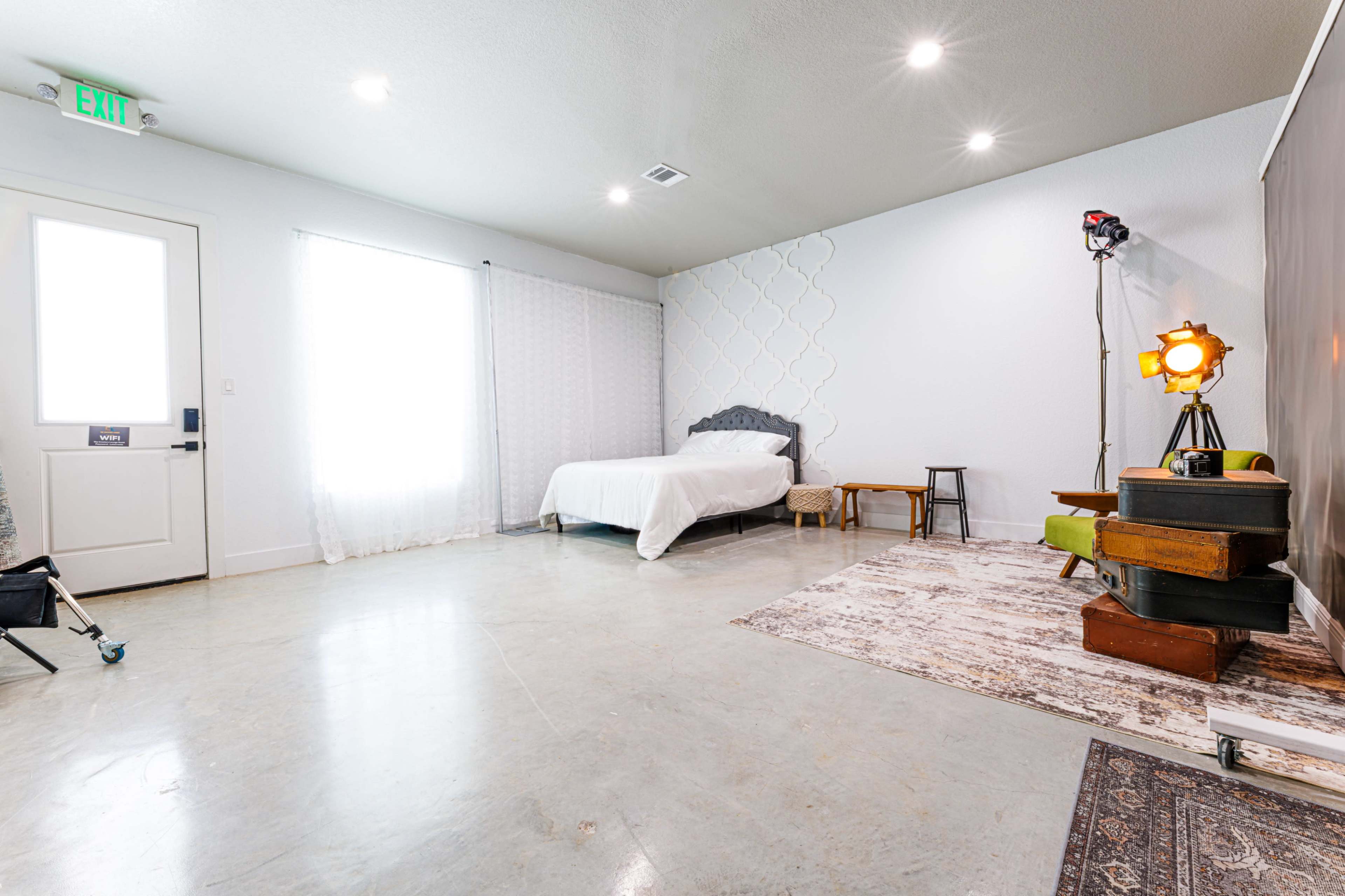 The image shows a minimalistic room featuring a bed against a wall, a small table, and a light stand, with large windows allowing natural light to enter.