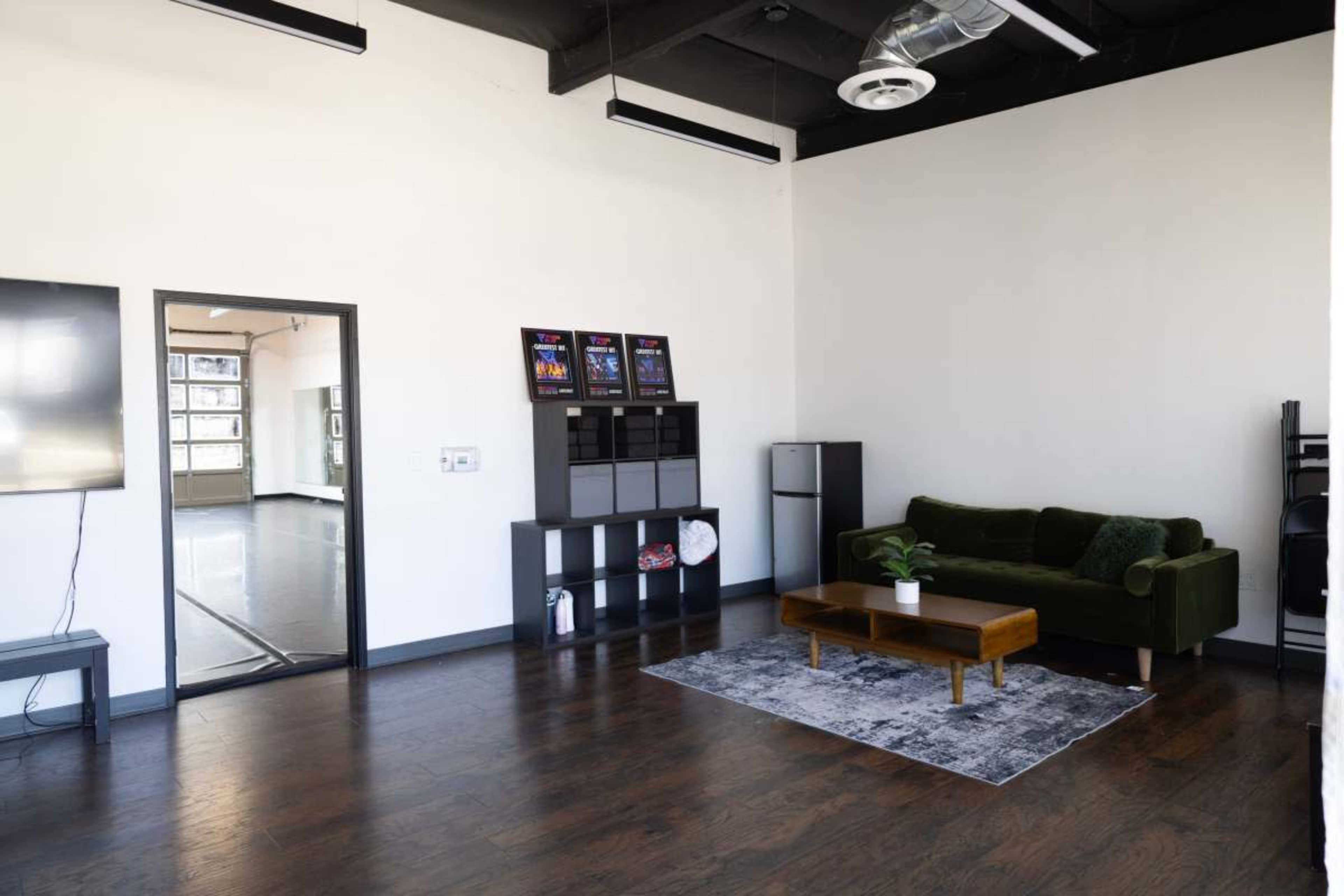 The image shows a spacious, minimalist interior with a green sofa, a wooden coffee table, and a decorative rug, along with shelves and a refrigerator in the background.