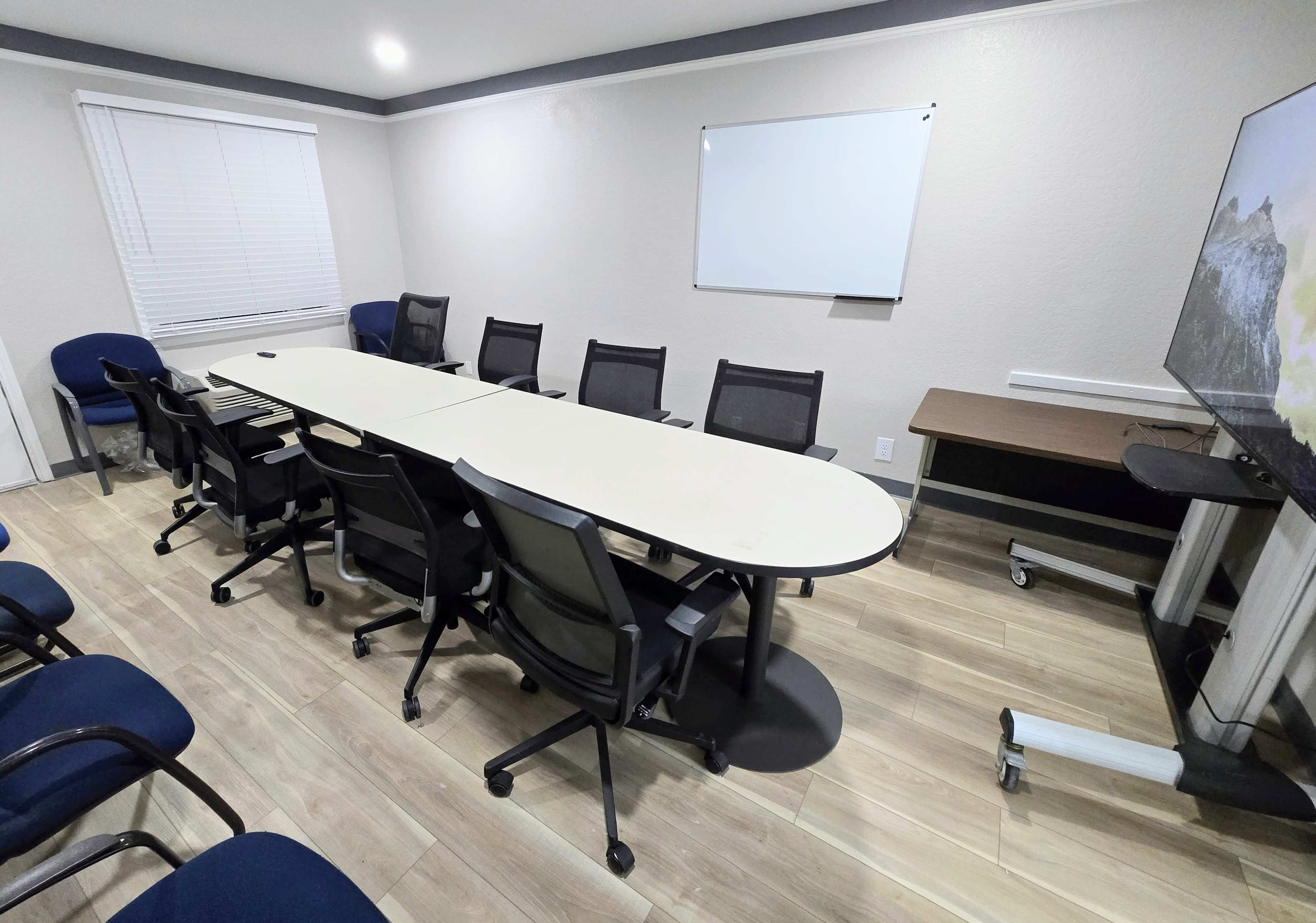 The image shows a conference room with a large oval table surrounded by chairs, a whiteboard on the wall, and a television mounted on a stand.