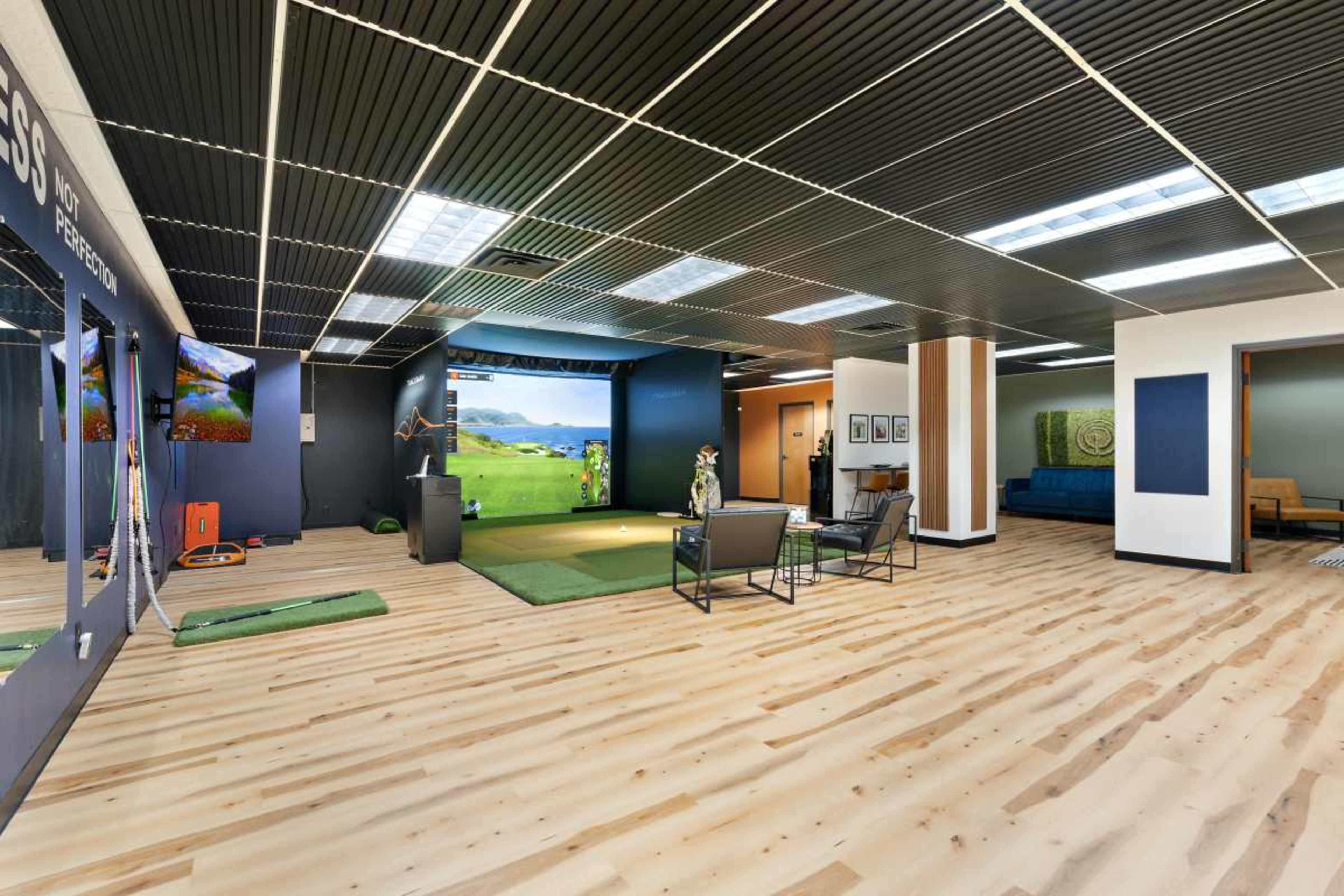 The image depicts a modern indoor golf simulation space featuring a large screen, turf area, seating, and multiple televisions mounted on the walls.