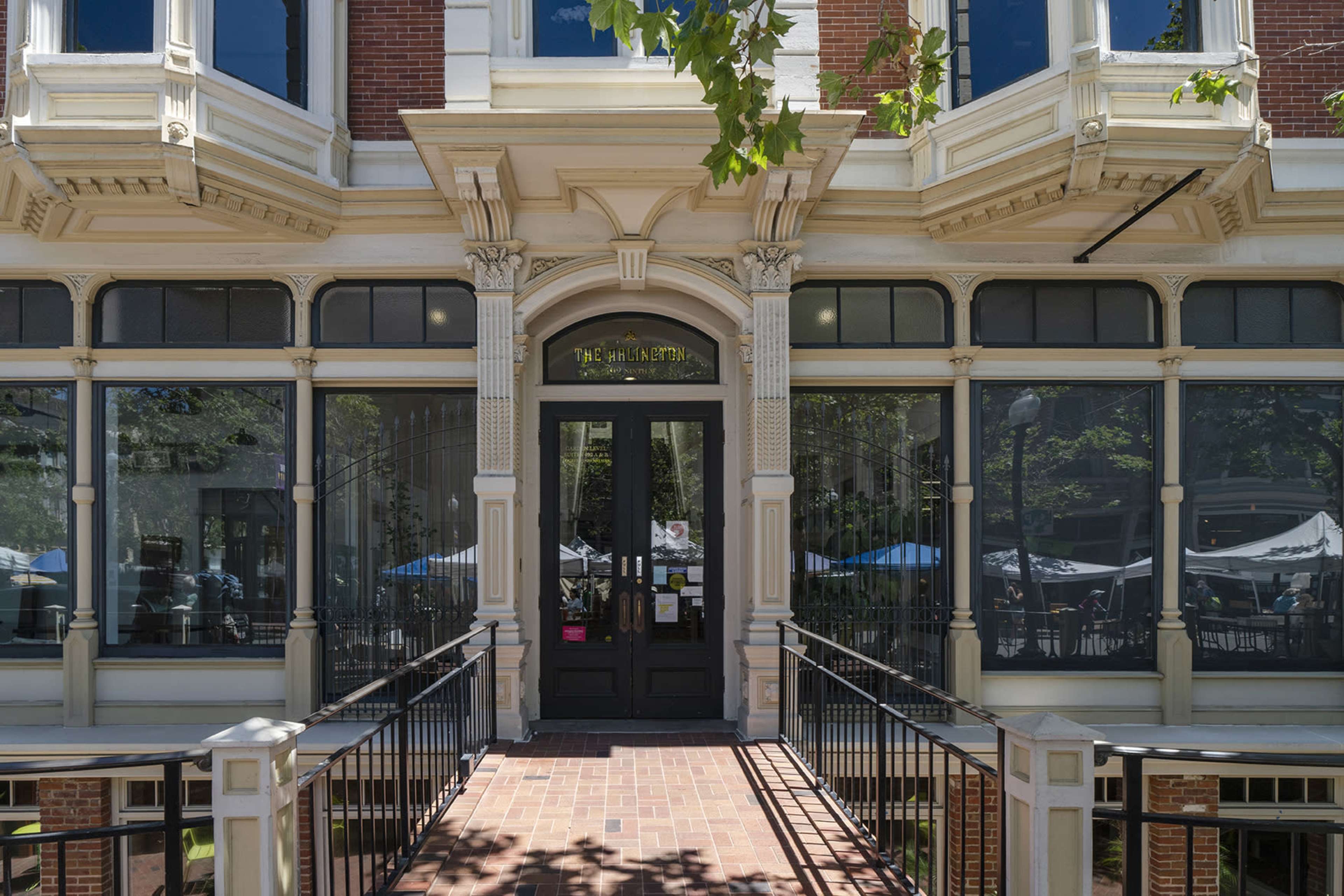 The entrance of a historic brick building with large windows and a decorative door, leading to an outdoor area with visible seating.