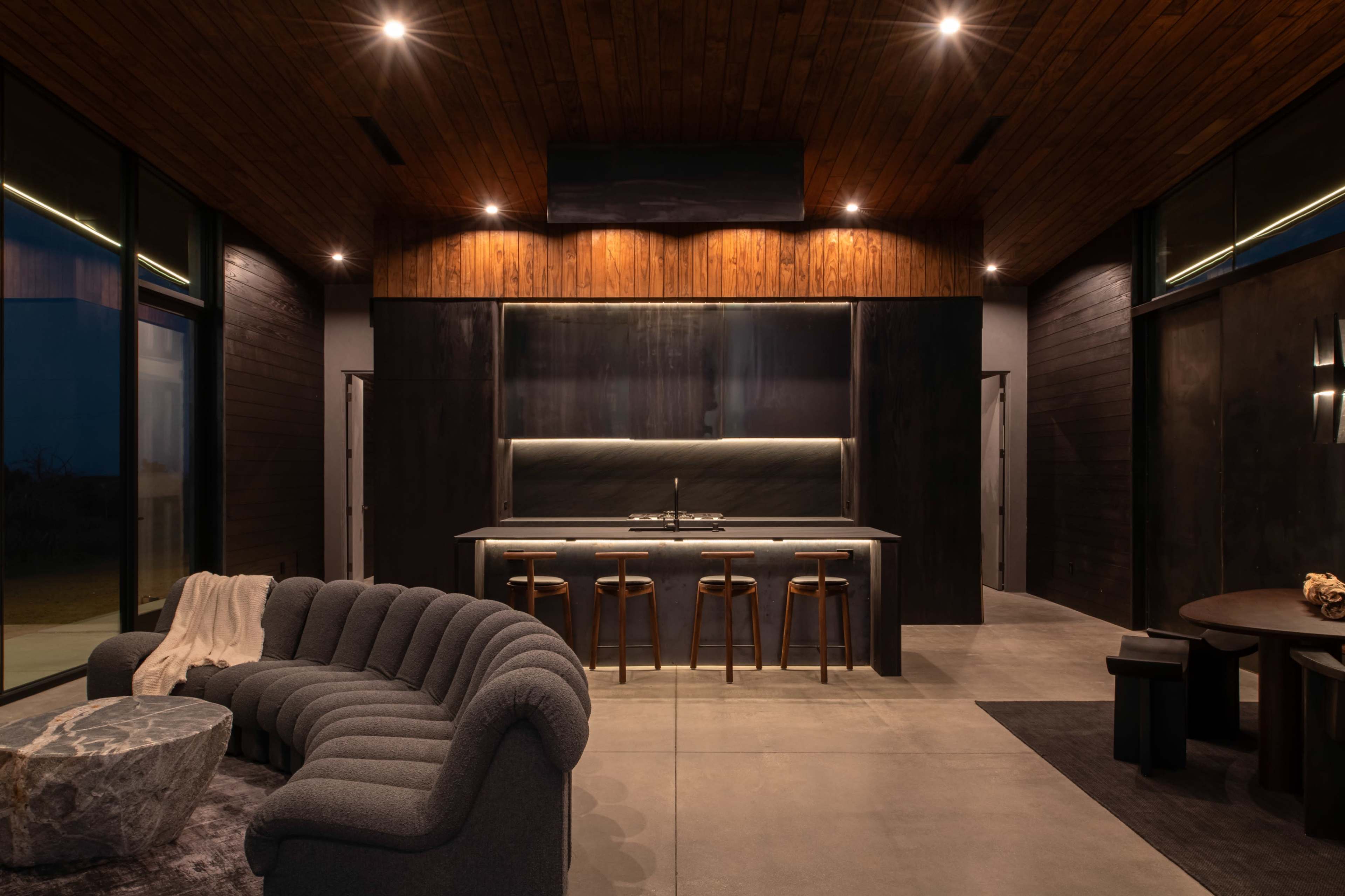 The image shows a modern interior space featuring a living area with a curved sofa, a kitchen with bar stools, and wood-paneled ceilings.