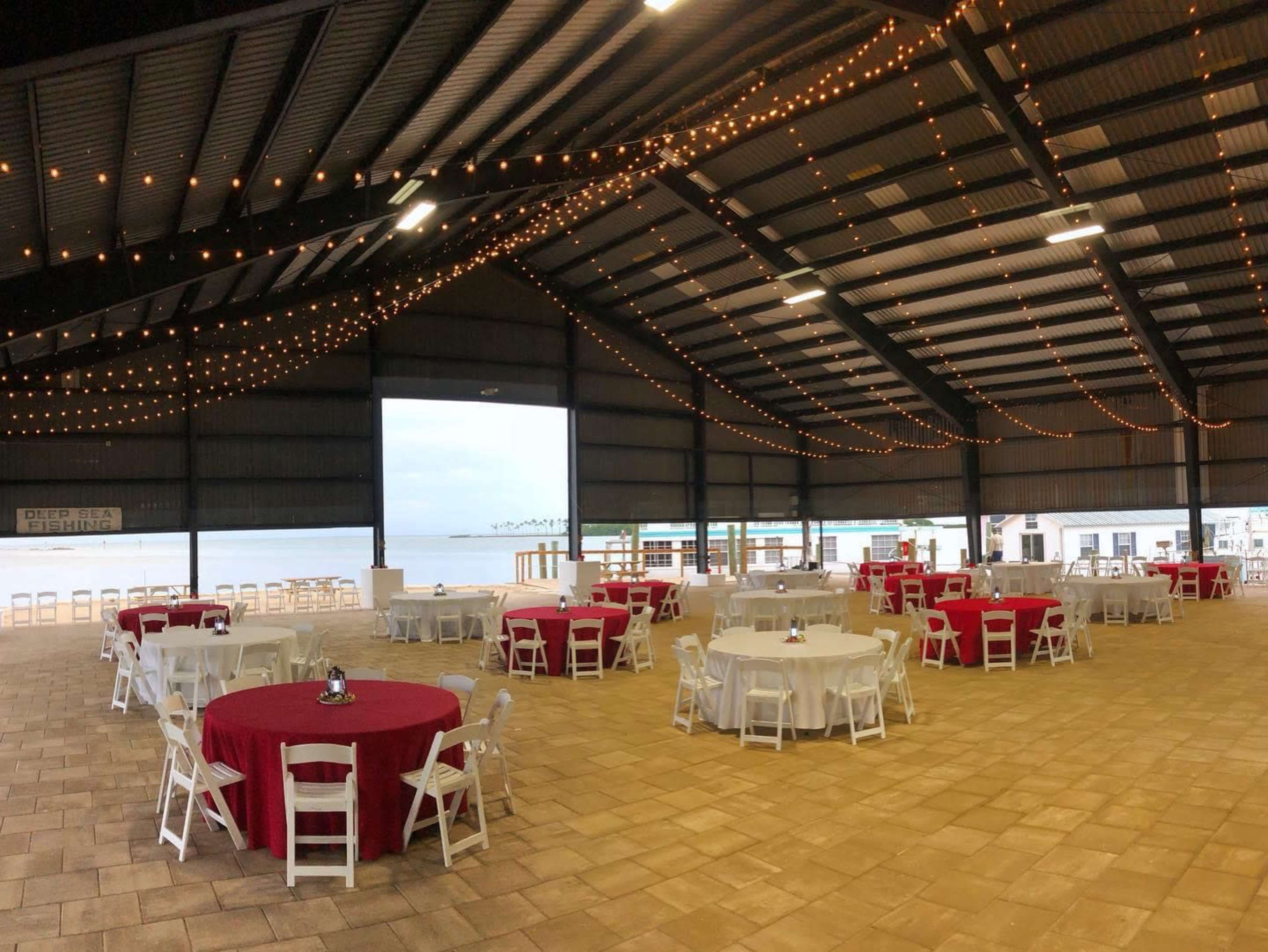 The image shows a spacious indoor event venue with round tables set up, featuring white tablecloths and red table runners, under warm string lights and a view of a waterfront through large openings.